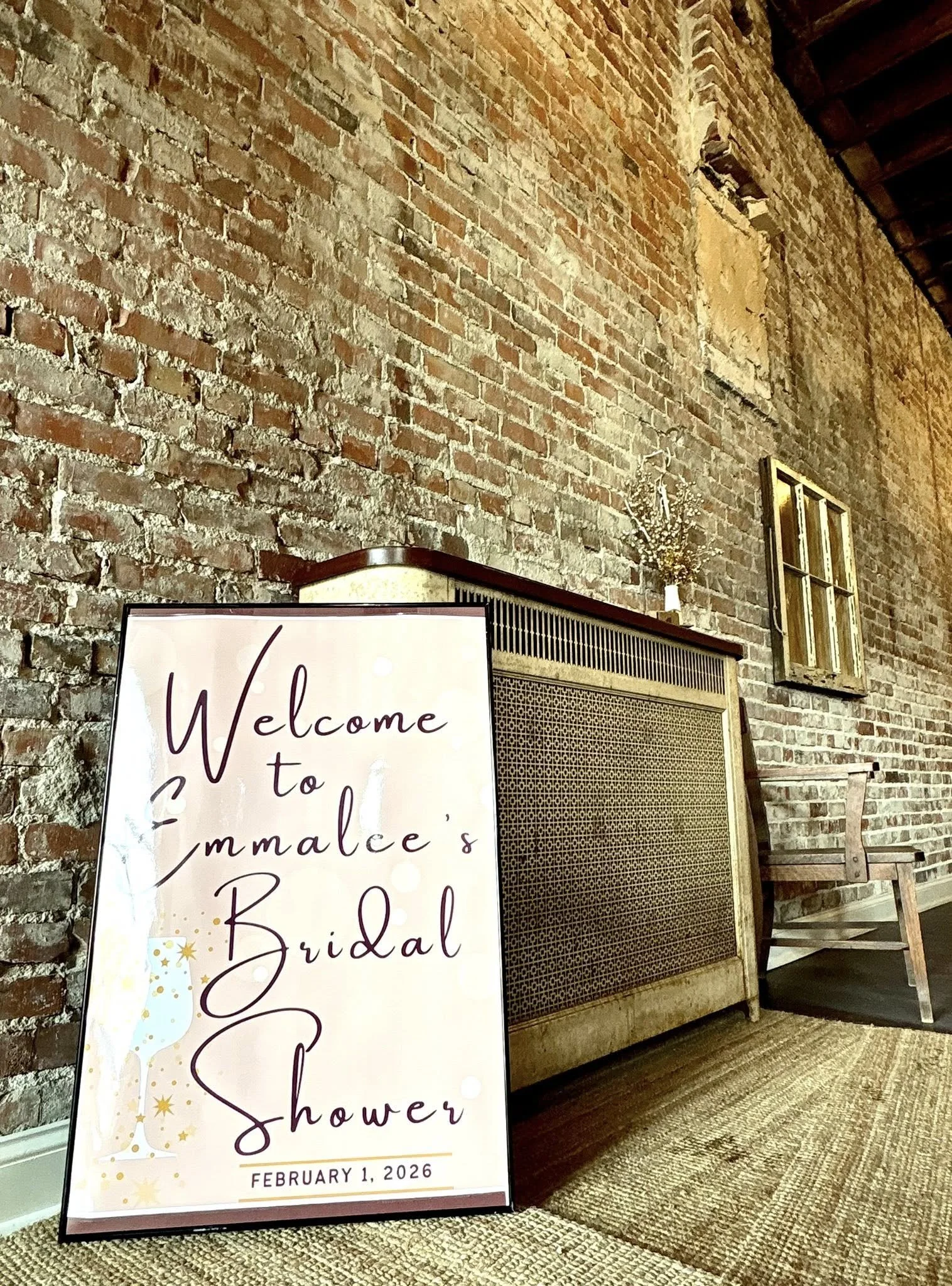 A welcome sign for Emmalee's bridal shower with an illustration of a champagne glass, placed on a textured floor against an exposed brick wall.