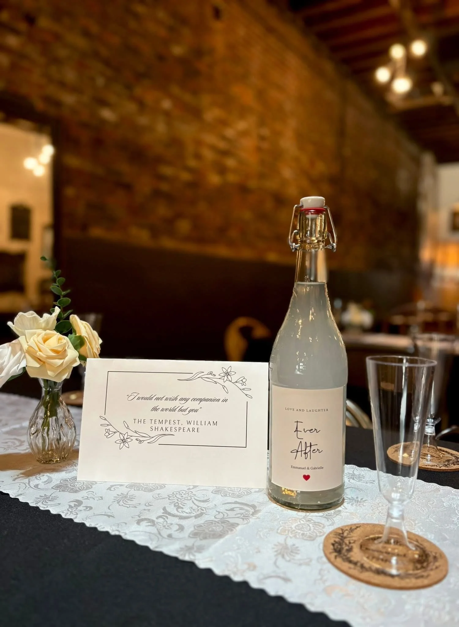A table setting with a small vase of white roses, a card with a quote from Shakespeare, a bottle of water labeled 'Ever After' with a heart, an empty glass on a coaster, and a brick wall in the background.