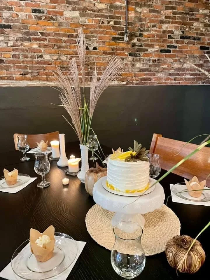 Decorated table with a white frosted cake on a cake stand, surrounded by candles, glassware, flower arrangements, and dessert cups in a dining setting with brick wall background.