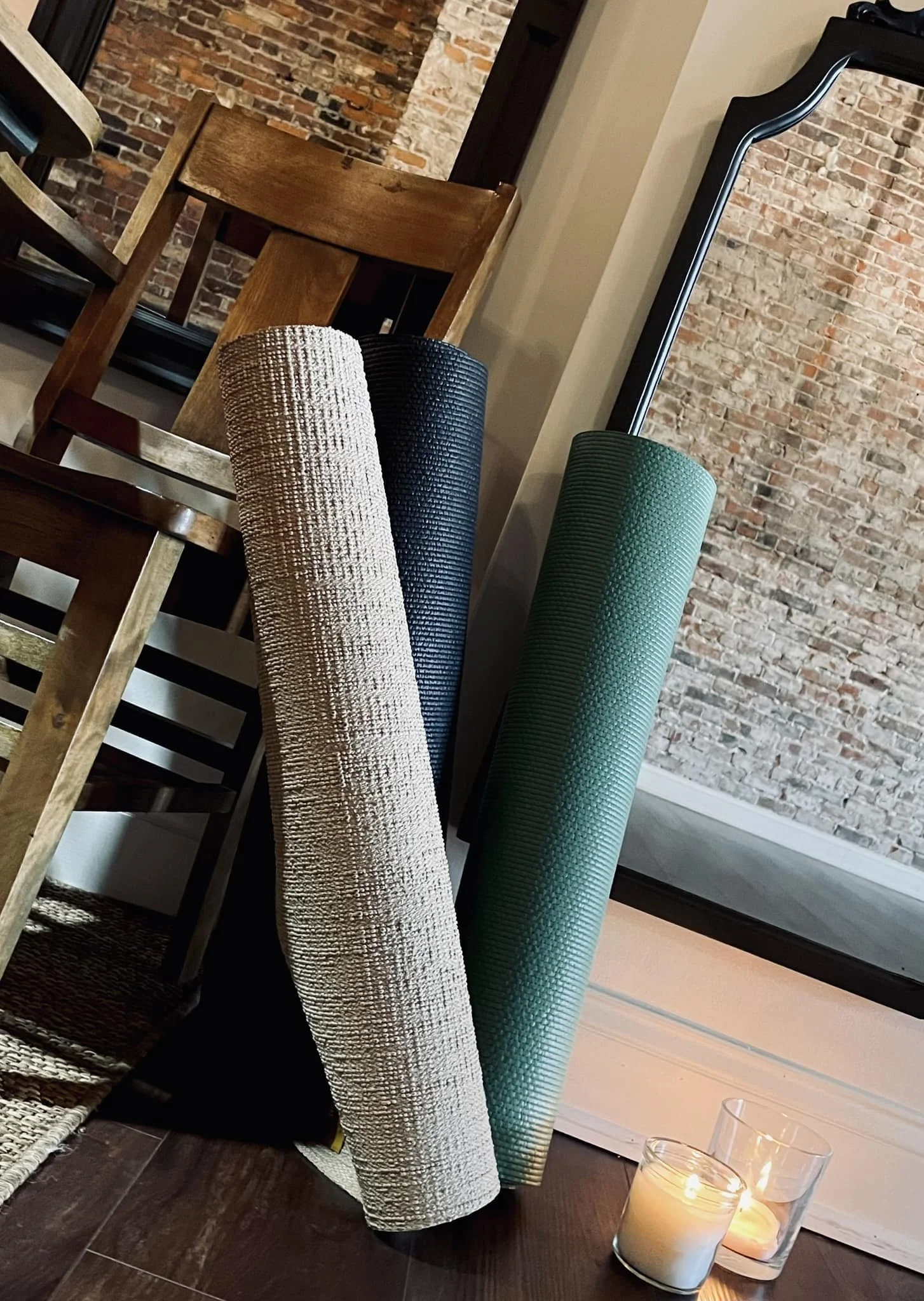 Four rolled-up yoga mats in beige, black, green, and gray colors leaning against a wall near candles on a dark wooden floor, with a brick wall and a mirror in the background.