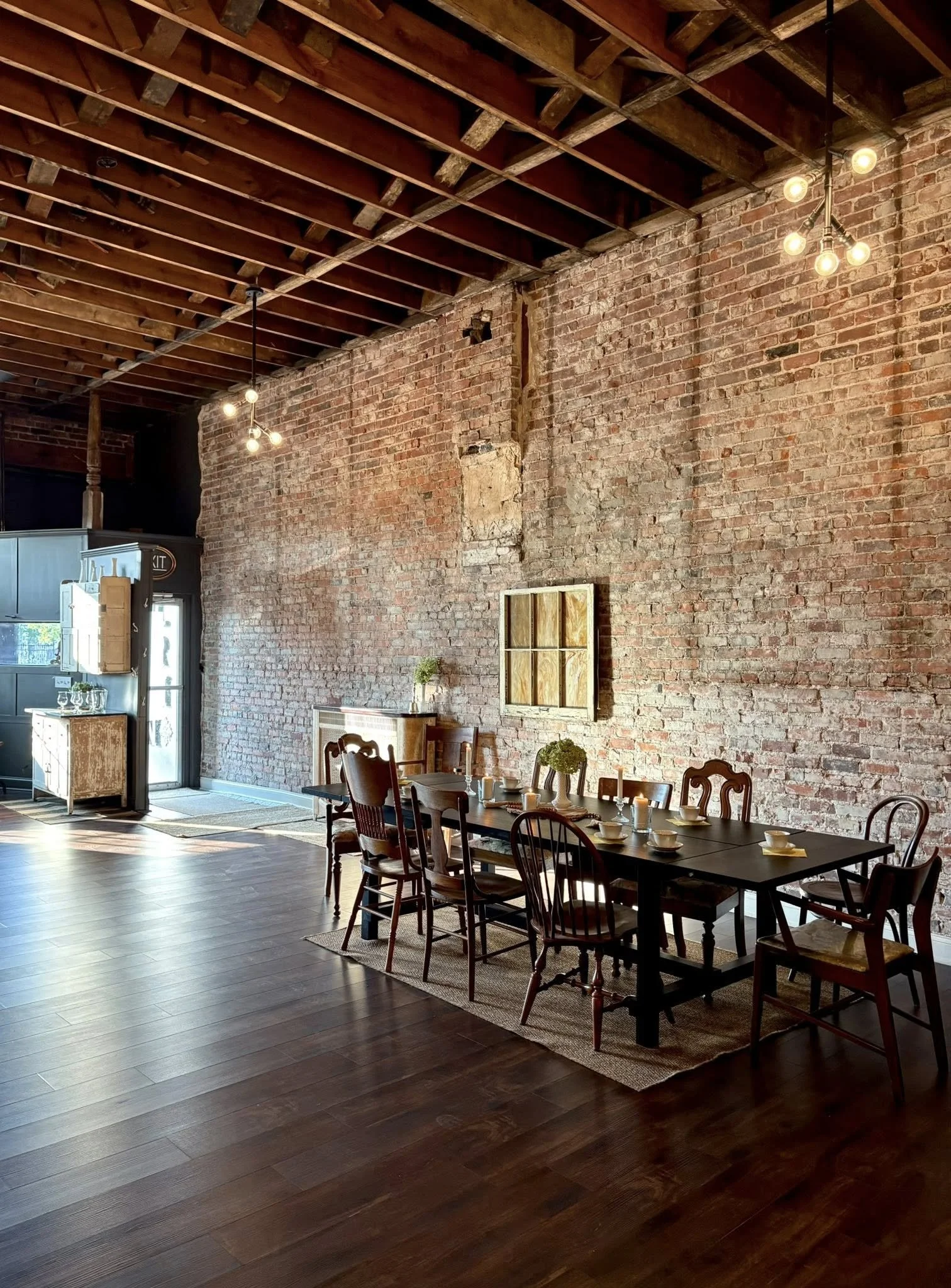 Interior of a rustic dining area with a long black table, wooden chairs, and an exposed brick wall. There are ceiling lights and a small window on the brick wall. The room has wooden flooring and minimal decor.