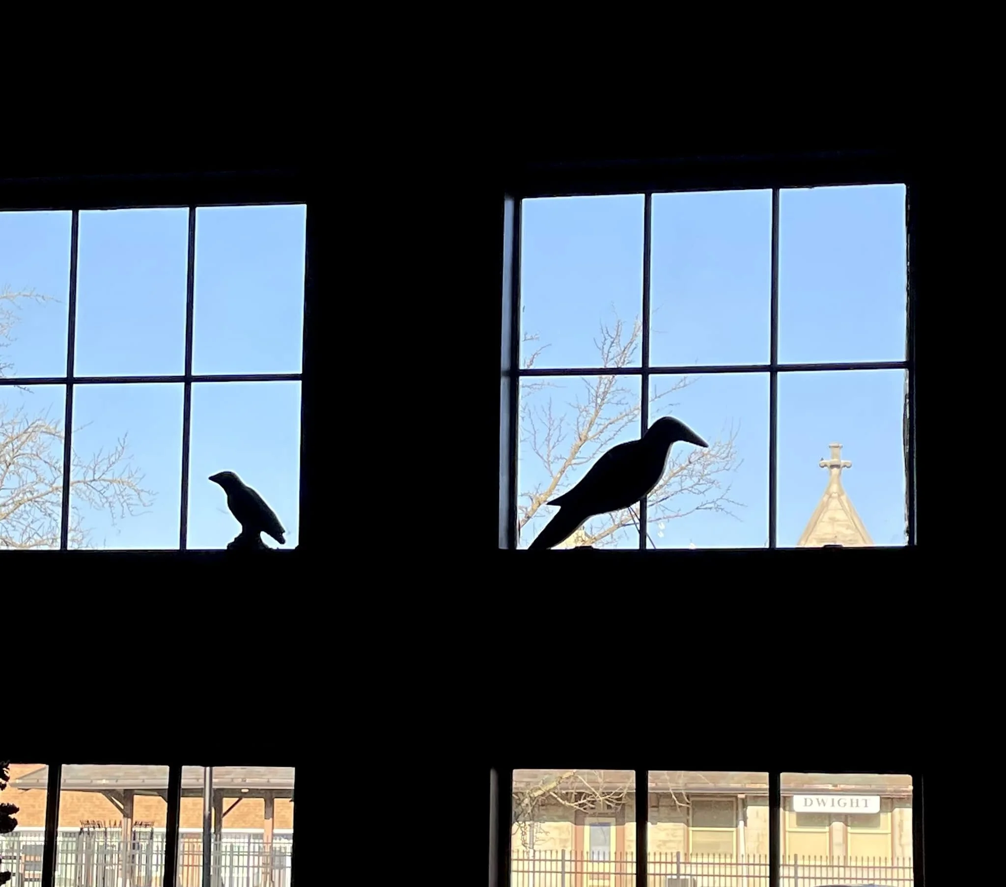 Silhouetted bird decorations on windowsill with view of clear blue sky, bare tree branches, and church steeple in the background.
