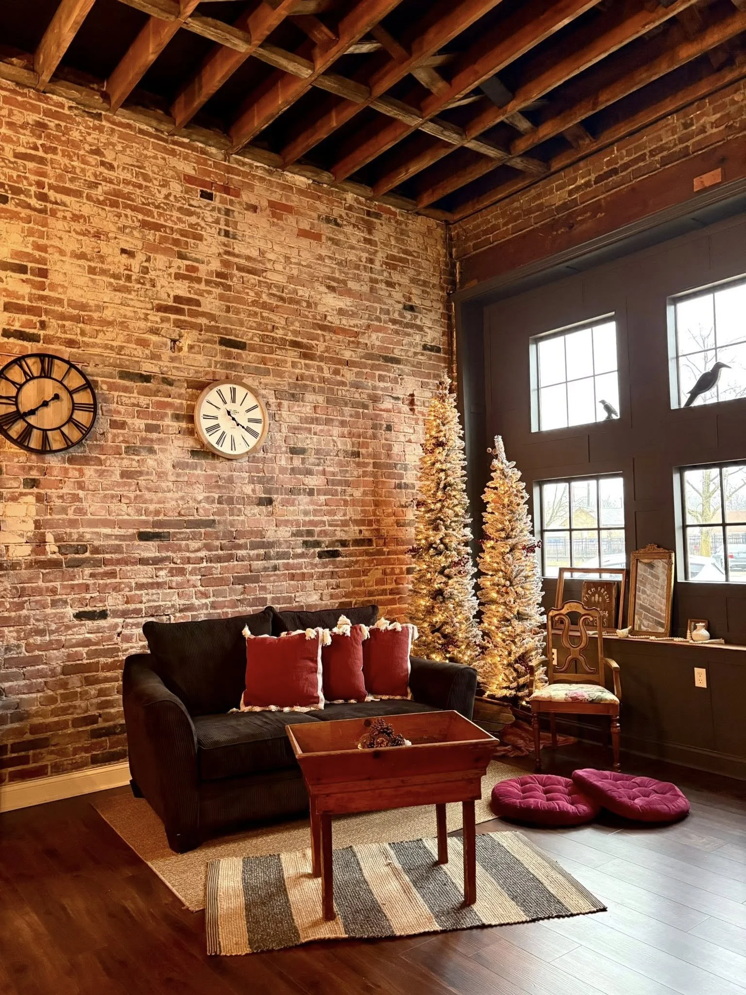 A cozy living room corner decorated for Christmas with a black sofa, red pillows, a small wooden table, two pink cushions, artificial Christmas trees, a large window with bird silhouettes, and exposed brick wall with two clocks.
