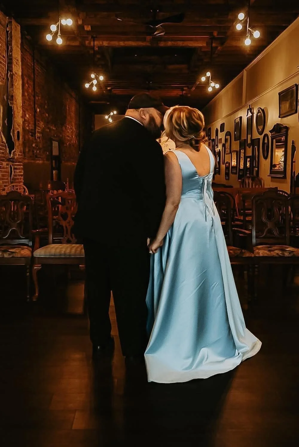 A couple dressed in formal attire sharing a private moment in a dimly lit restaurant or event space with exposed brick walls and vintage furniture.