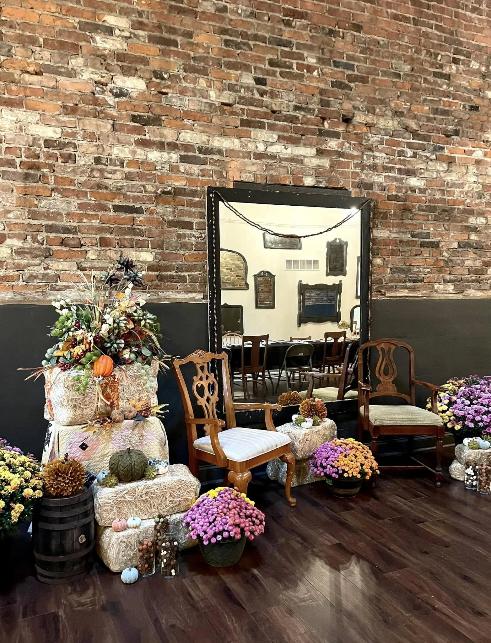 Indoor space with a brick wall, decorated with chairs, potted flowers, pumpkins, and fall decorative items, with a large mirror reflecting part of the room.