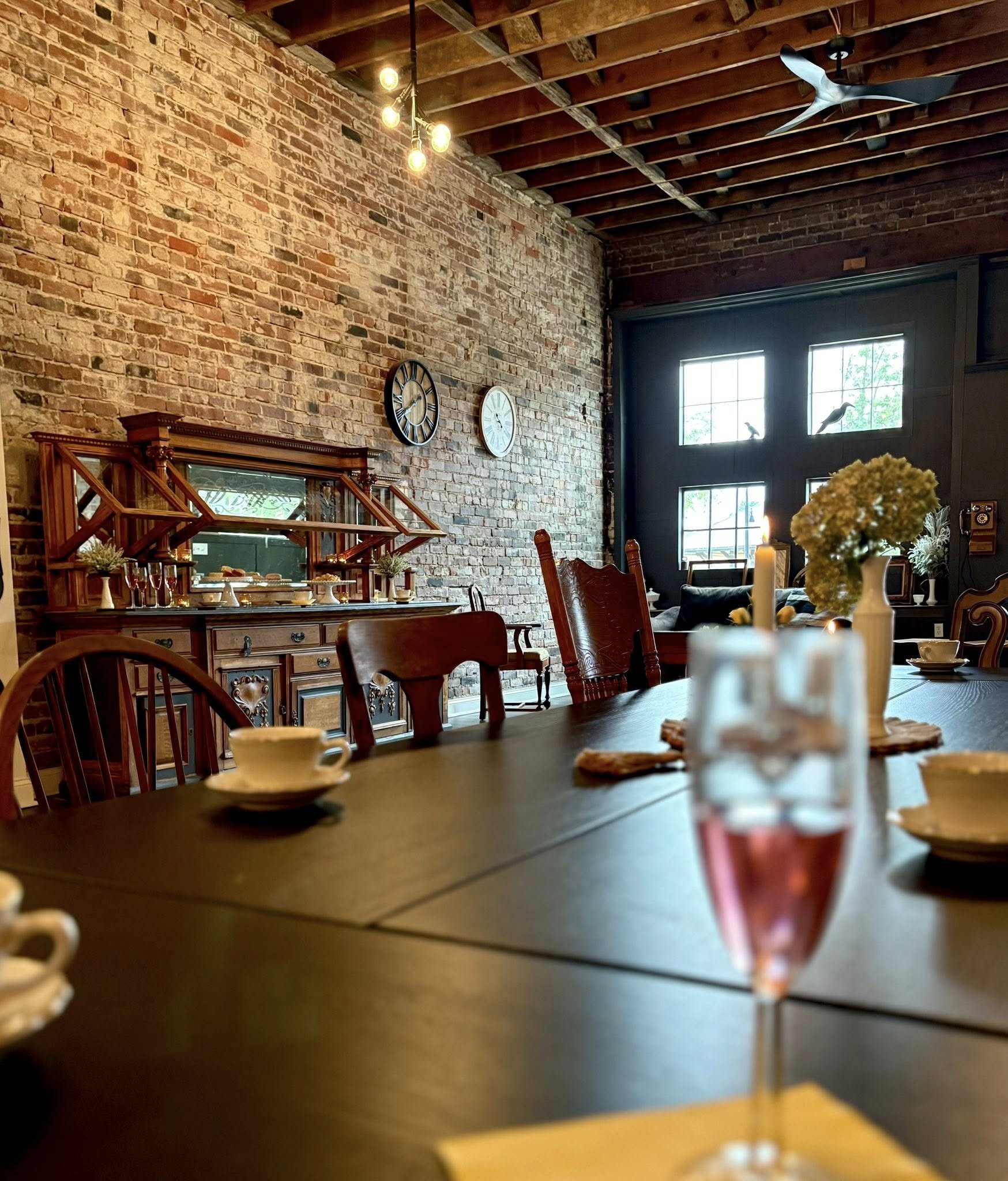 Interior of a rustic dining room with a brick wall, wooden ceiling, and large windows. The table is set with cups, bowls, and a glass of pink drink, with a floral centerpiece and candle. Antique furniture and clocks are visible.