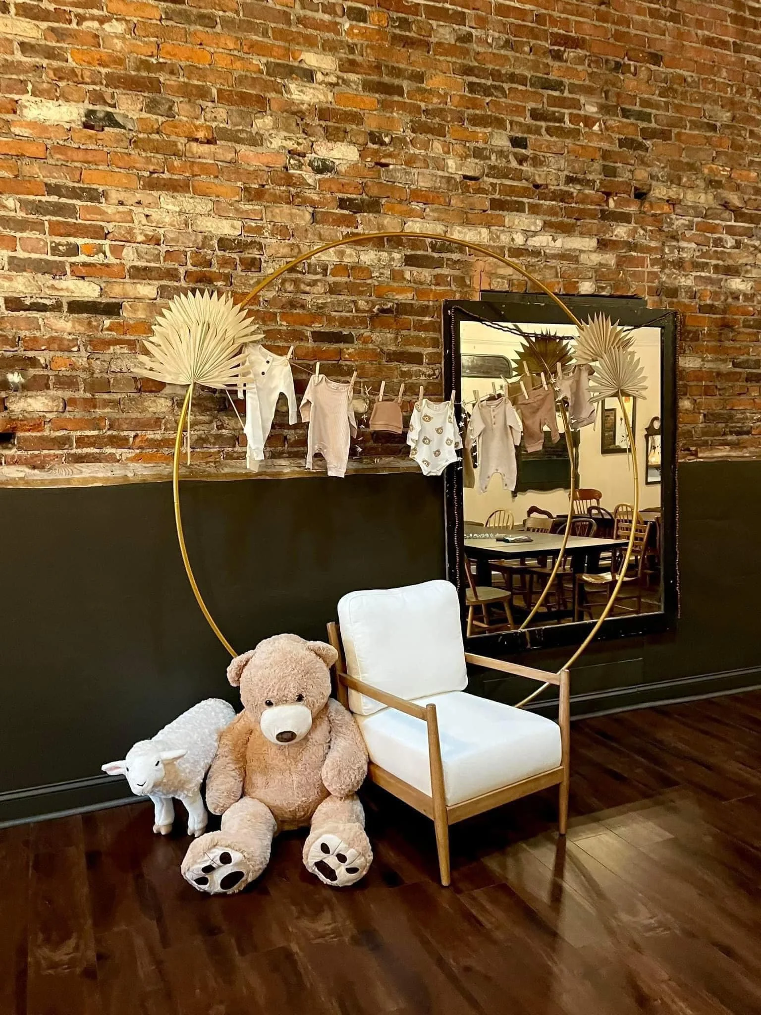 Toys including a large teddy bear, a small plush sheep, and a plush lamb, positioned next to a white armchair, with a brick wall backdrop decorated with a mirror, hanging clothes, and dried palm leaves.