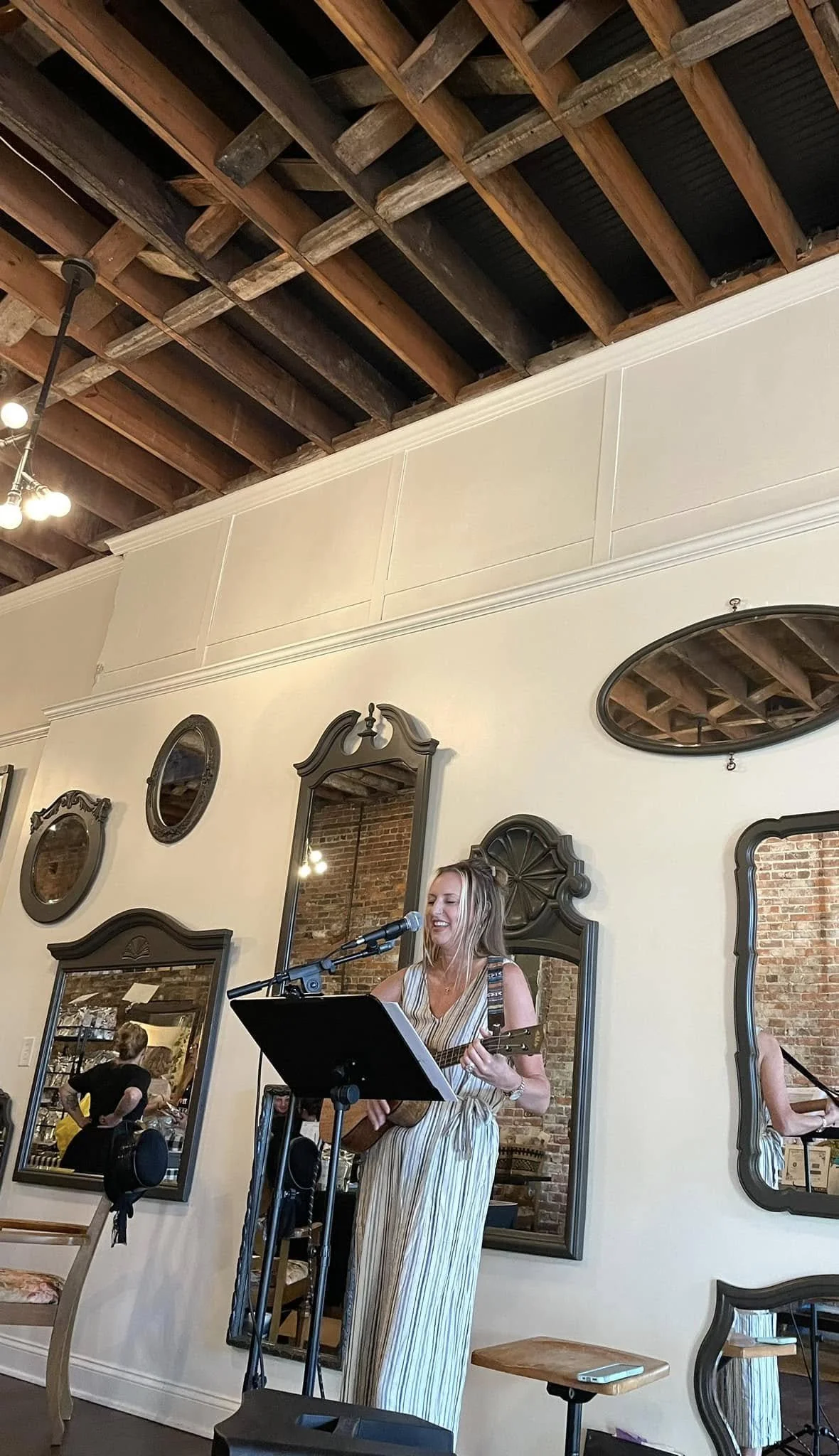 A woman in a striped dress singing and playing guitar with a microphone in a cafe or restaurant. The background has a cream-colored wall with multiple mirrors and a visible brick wall reflection.