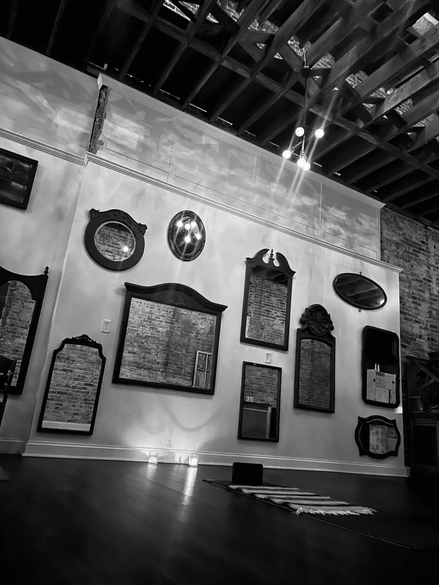 Interior of a room with a wall decorated with various vintage mirrors and frames, dim candle lighting, exposed brick walls, and a wooden ceiling.