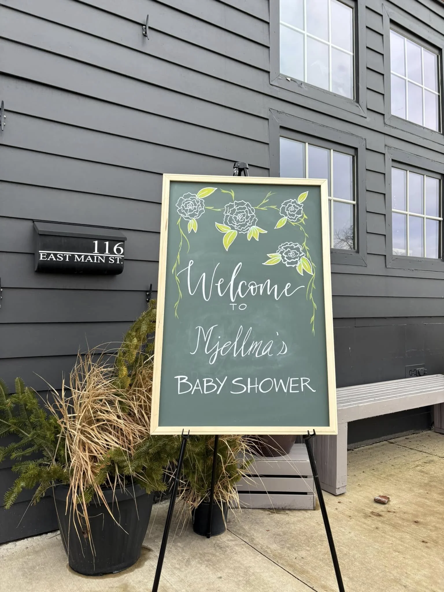 A welcome sign on a black easel outside a grey building. The sign reads 'Welcome to Myellma's Baby Shower' with decorative flowers at the top. There are plants in pots nearby, and a bench is to the right.
