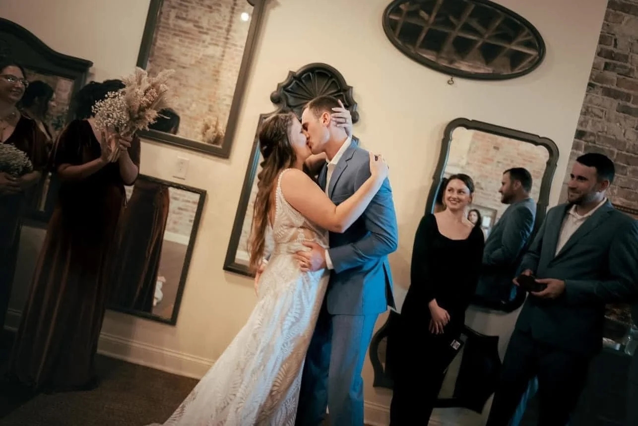 A couple in wedding attire sharing a kiss in a room with friends and family, with women holding bouquets and men in suits, decorated with large wall mirrors and exposed brick walls.