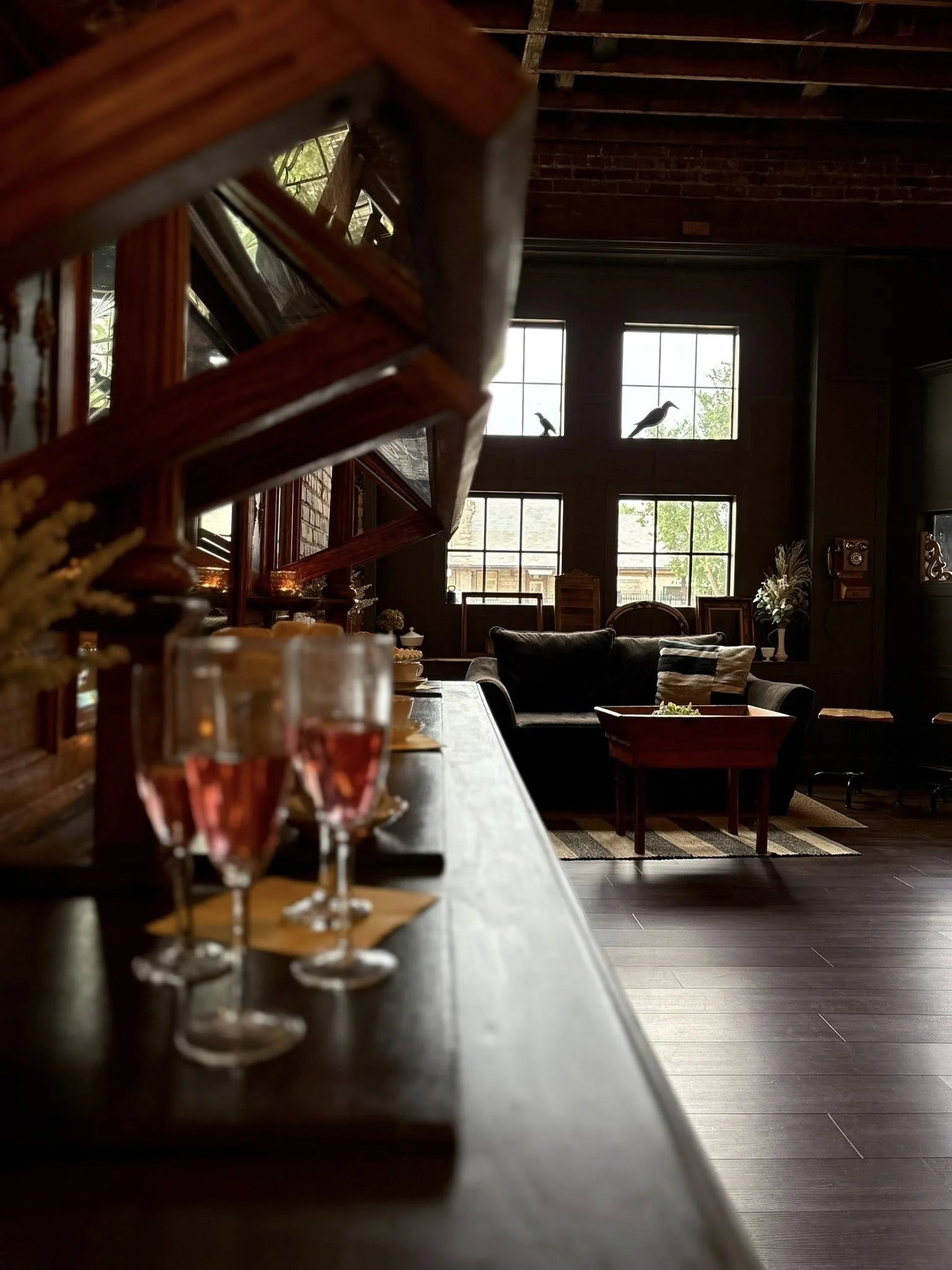 Inside a cozy room with large windows, a bar counter with glasses of pink drink, a black sofa, and decorative birds on the upper window. Natural light streams through the windows.