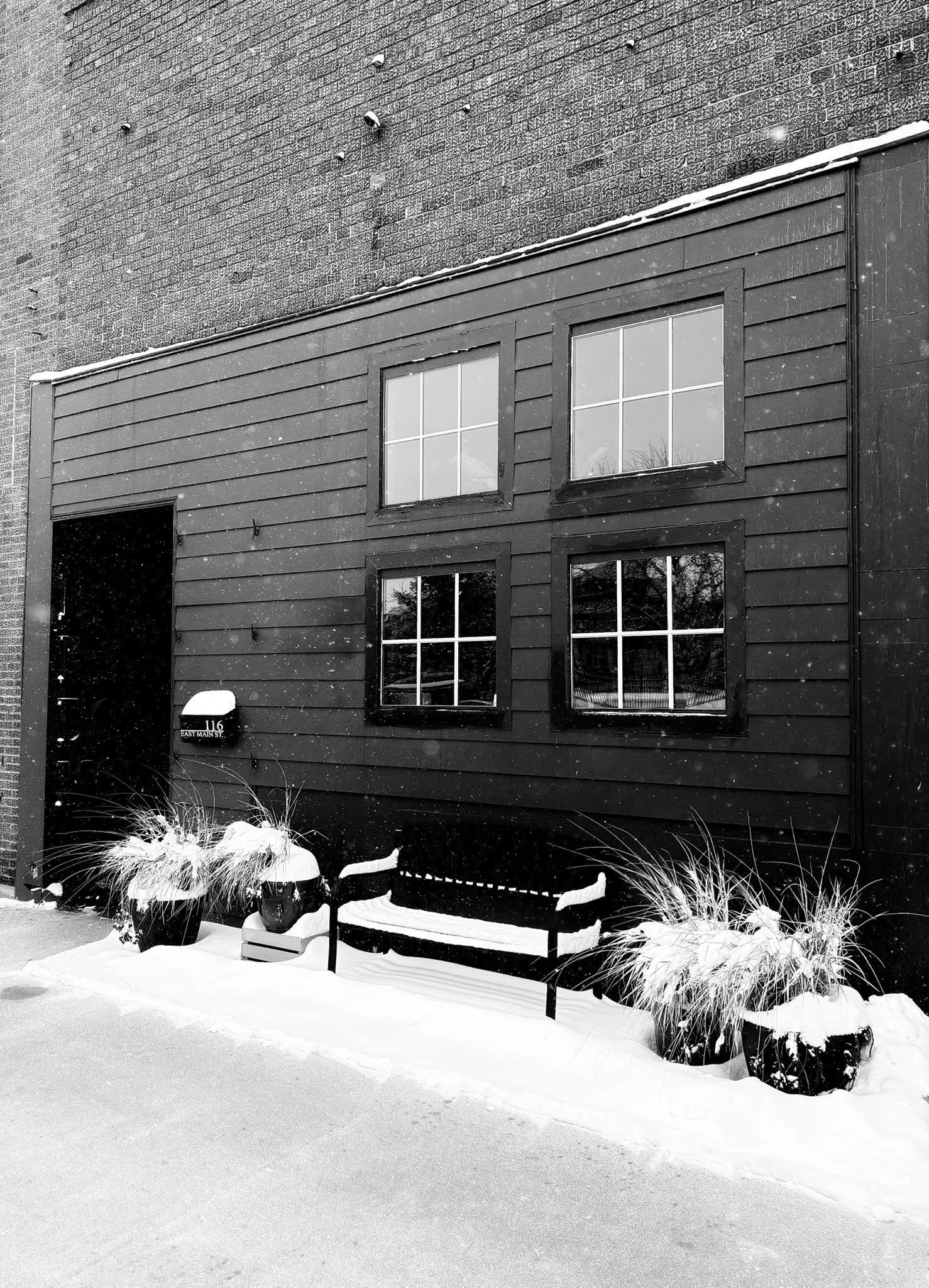 Black building with wooden siding, four windows, snow on the ground, potted plants, and a bench outside.