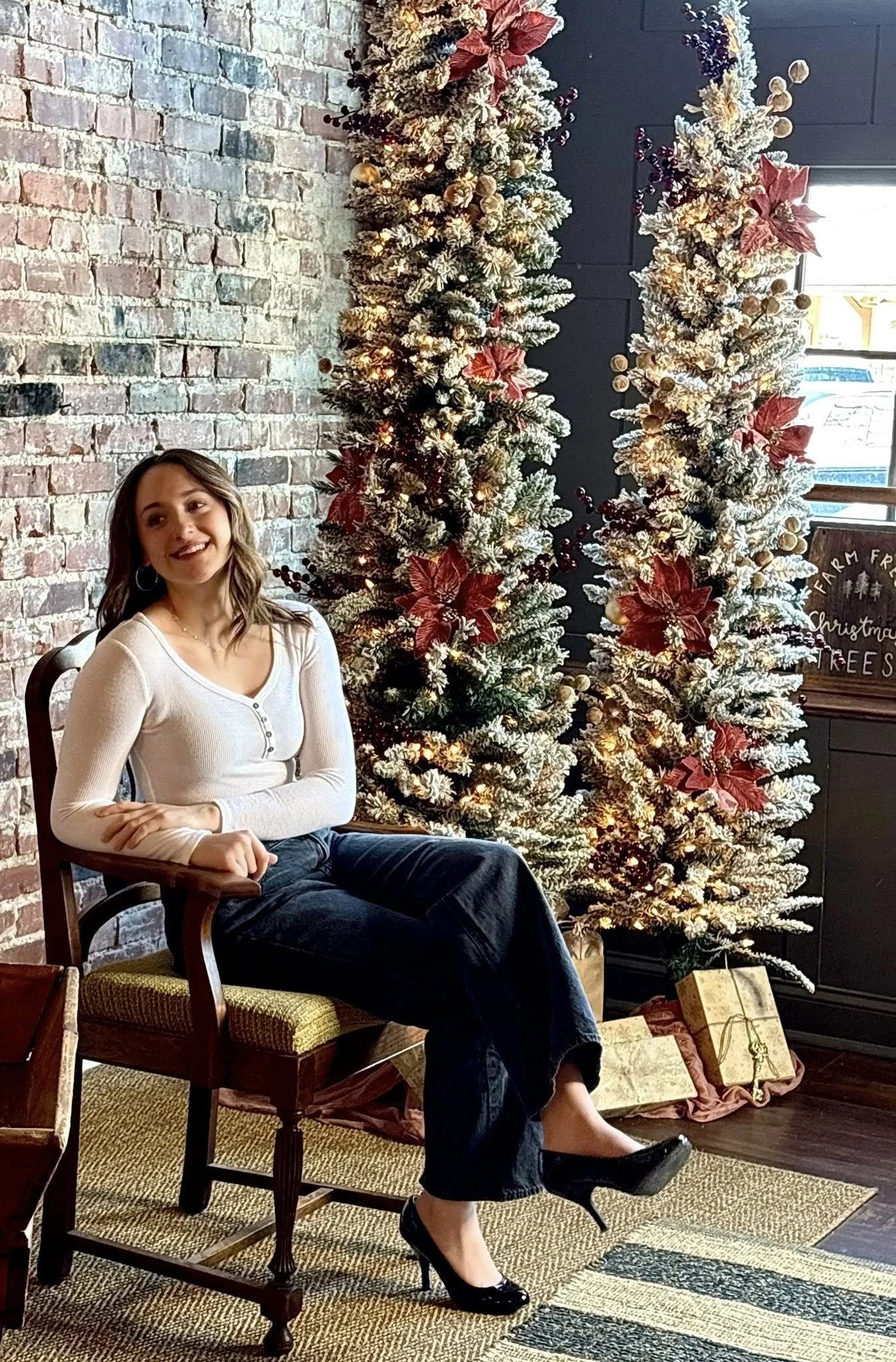 A woman sits on a wooden chair next to two decorated Christmas trees with poinsettia and pinecone ornaments, in a cozy indoor setting with brick and dark walls.