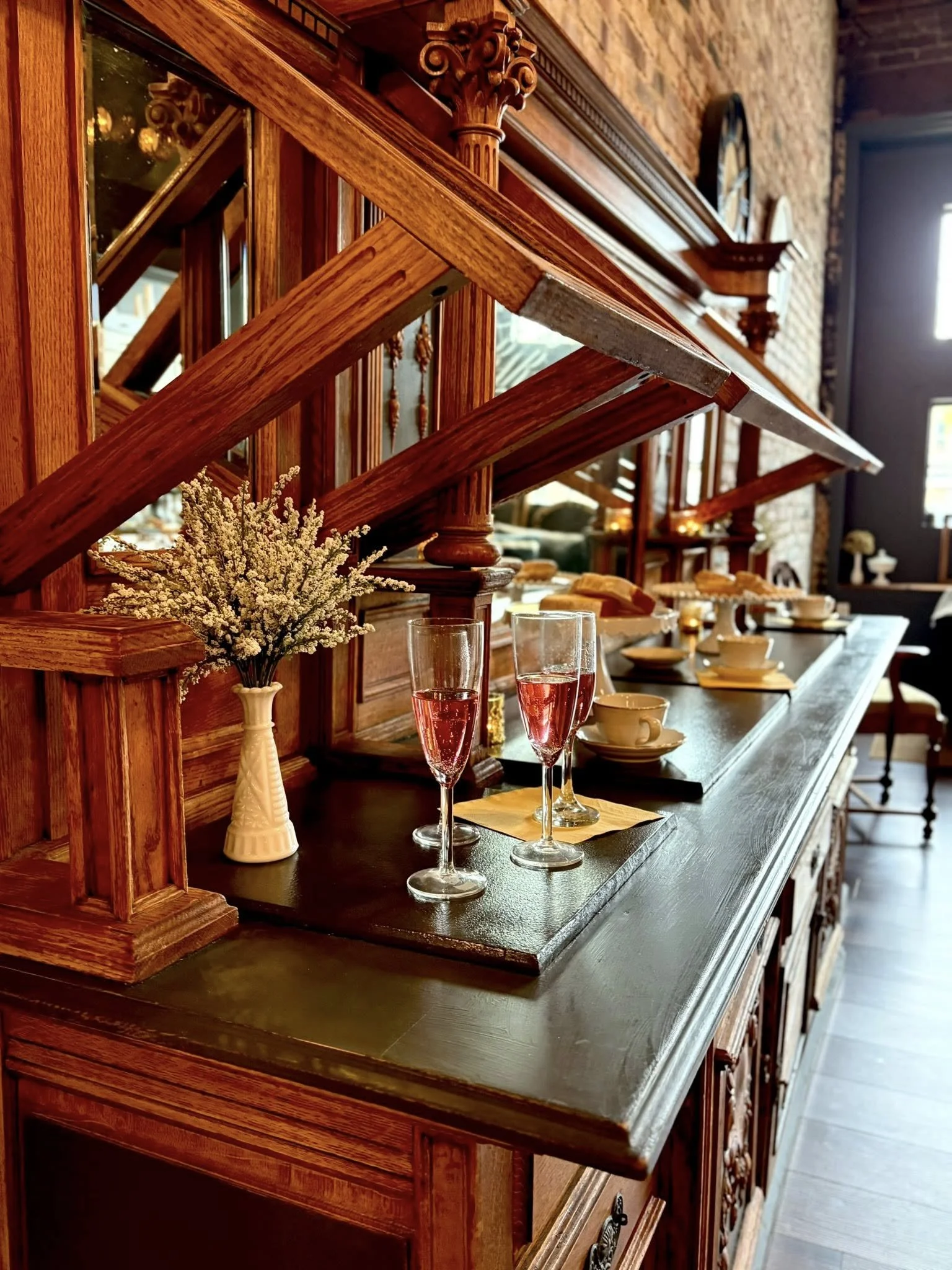 Dining room with a long wooden sideboard, set with cups, glasses, and a flower arrangement, featuring brick walls and large windows.