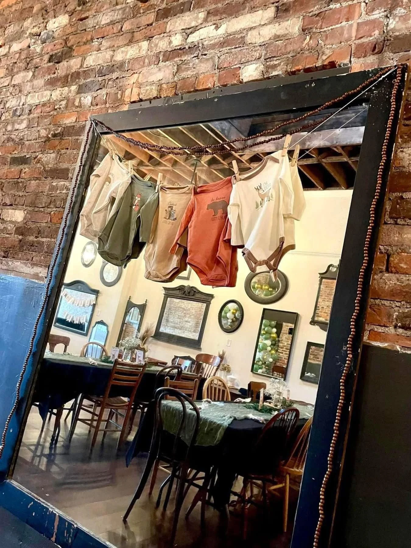 A mirror reflecting a decorated dining room with tables, chairs, and wall art, with a clothesline of baby onesies hanging above the mirror.