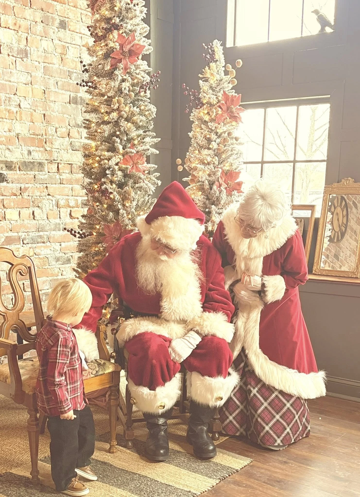 A young boy with blonde hair in a plaid red shirt and dark pants stands near Santa Claus and Mrs. Claus during a Christmas event. Santa is seated, dressed in a red suit with white fur trim, and Mrs. Claus is leaning over, wearing a red dress with whi