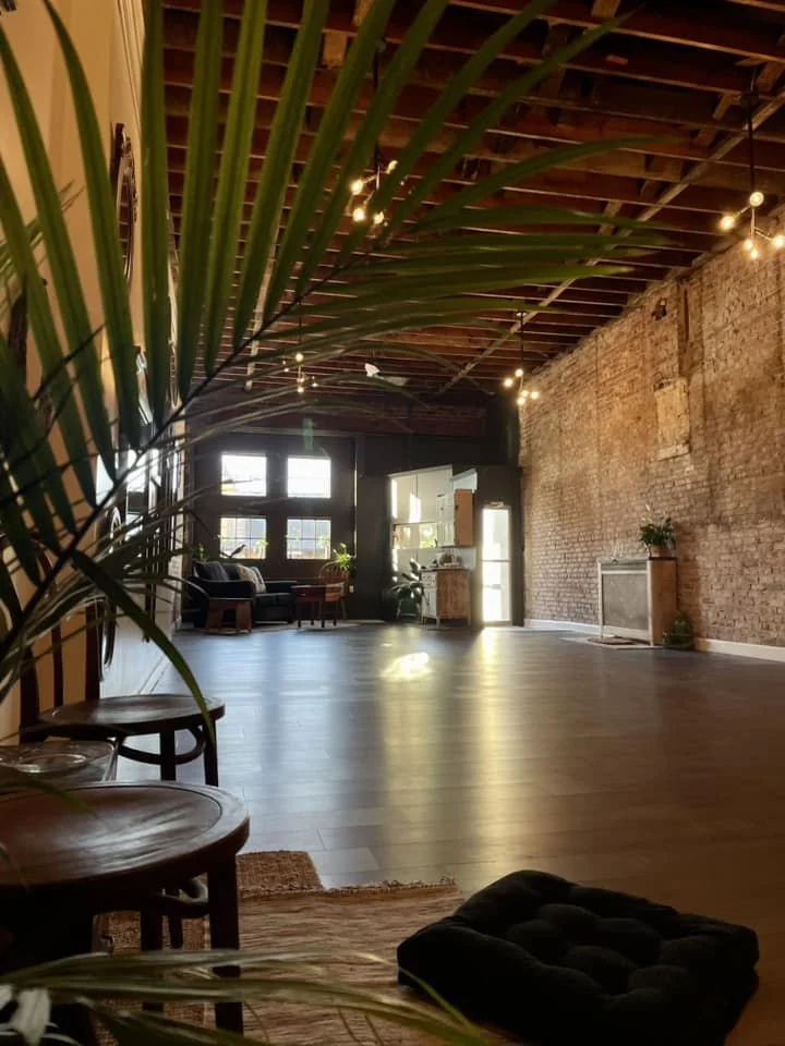 Spacious, softly lit living room with exposed brick wall, large windows, hardwood floors, and some furniture in the background, viewed partly through a palm plant in the foreground.