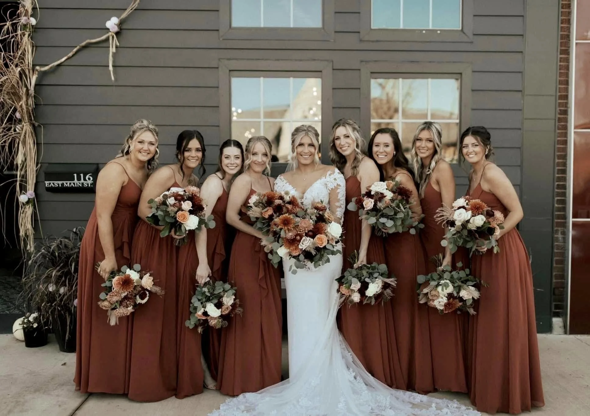 A bride and eight bridesmaids posing outside in front of a dark gray building with large windows. The bride is wearing a white lace wedding dress, and the bridesmaids are wearing matching long rust-colored dresses. They are all holding bouquets of fl