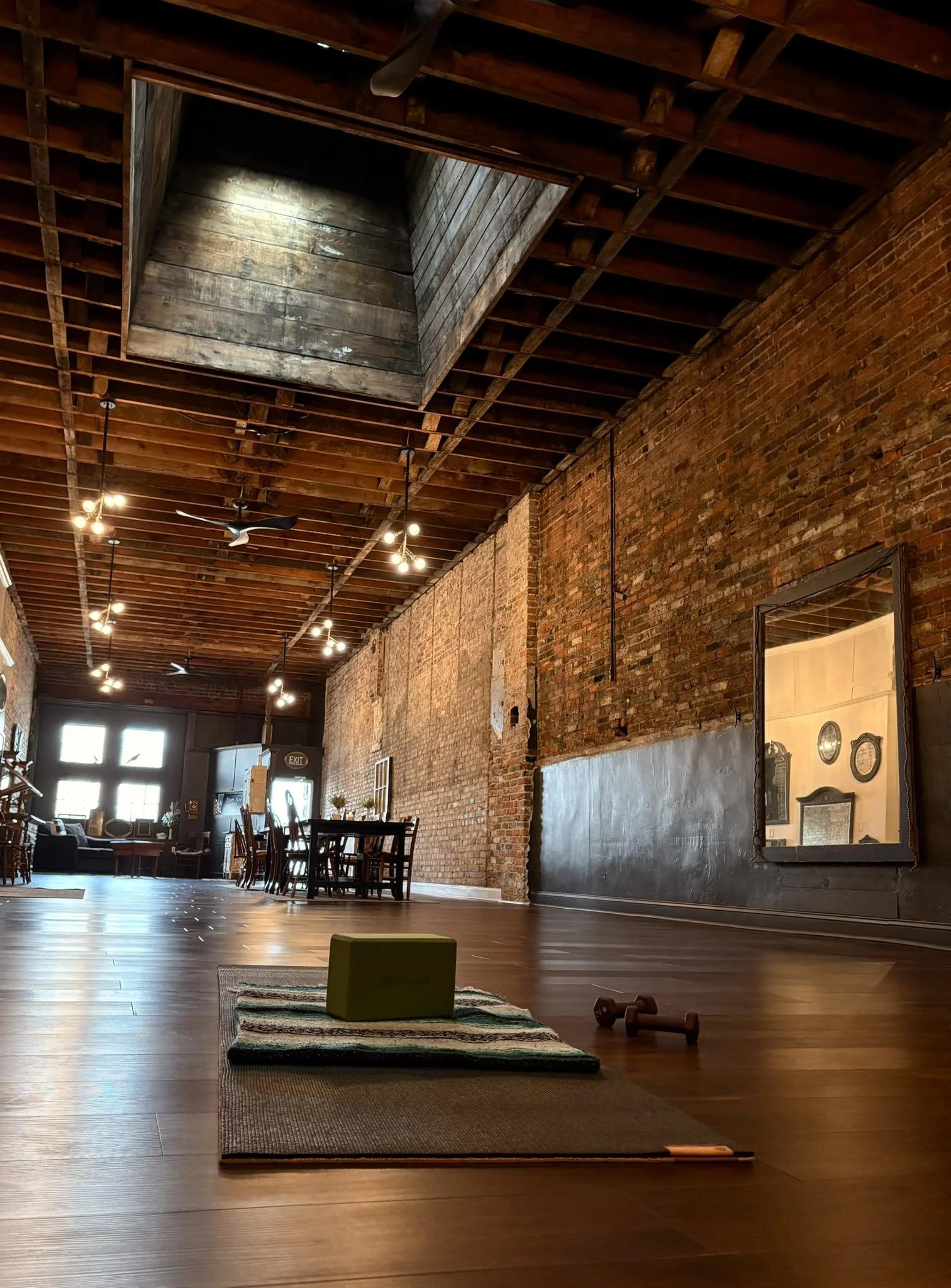 An empty loft-style room with wooden floors, exposed brick walls, and a wooden ceiling. A yoga mat, a green yoga block, and a small dumbbell are on the floor in the foreground. Sunlight streams in through large windows at the back, and there are tabl