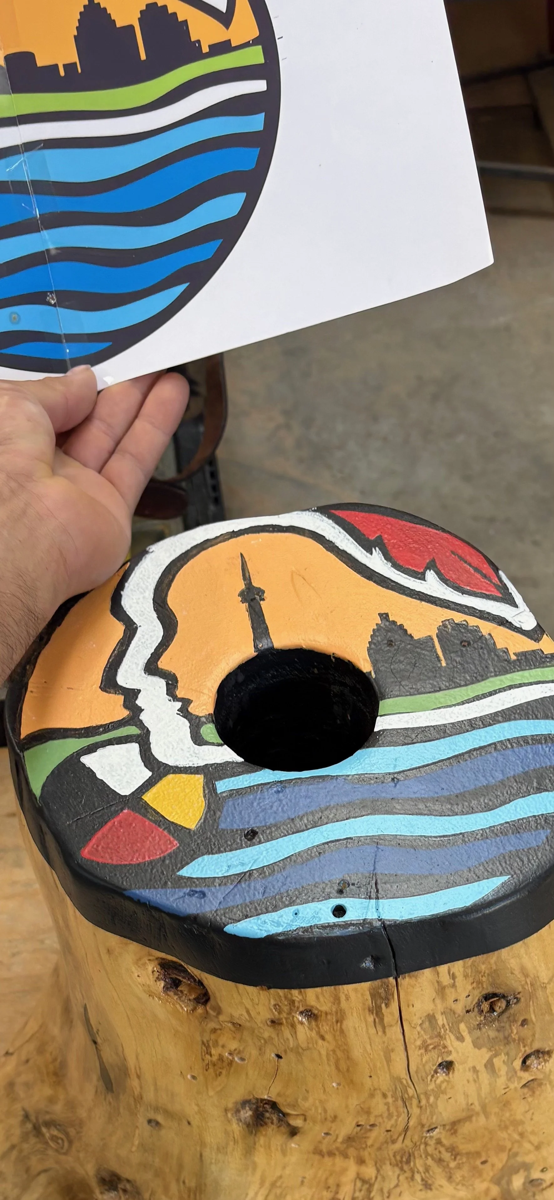 A painted wooden stool with a cityscape and water design, featuring an orange background with a silhouette of the CN Tower, and a piece of paper with a similar design being held above it.