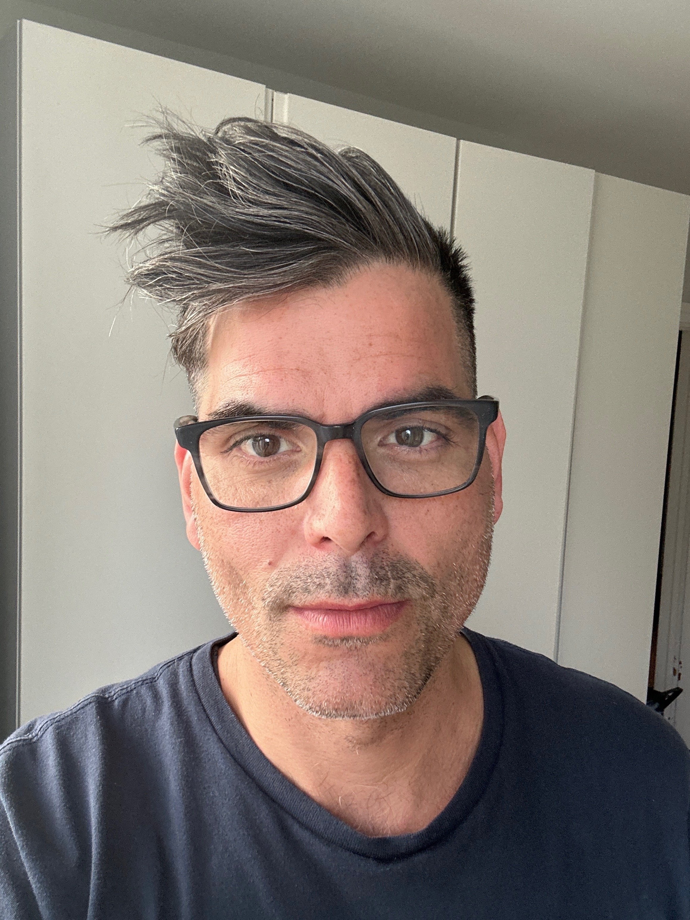 A man with styled hair, glasses, and facial stubble taking a selfie indoors.