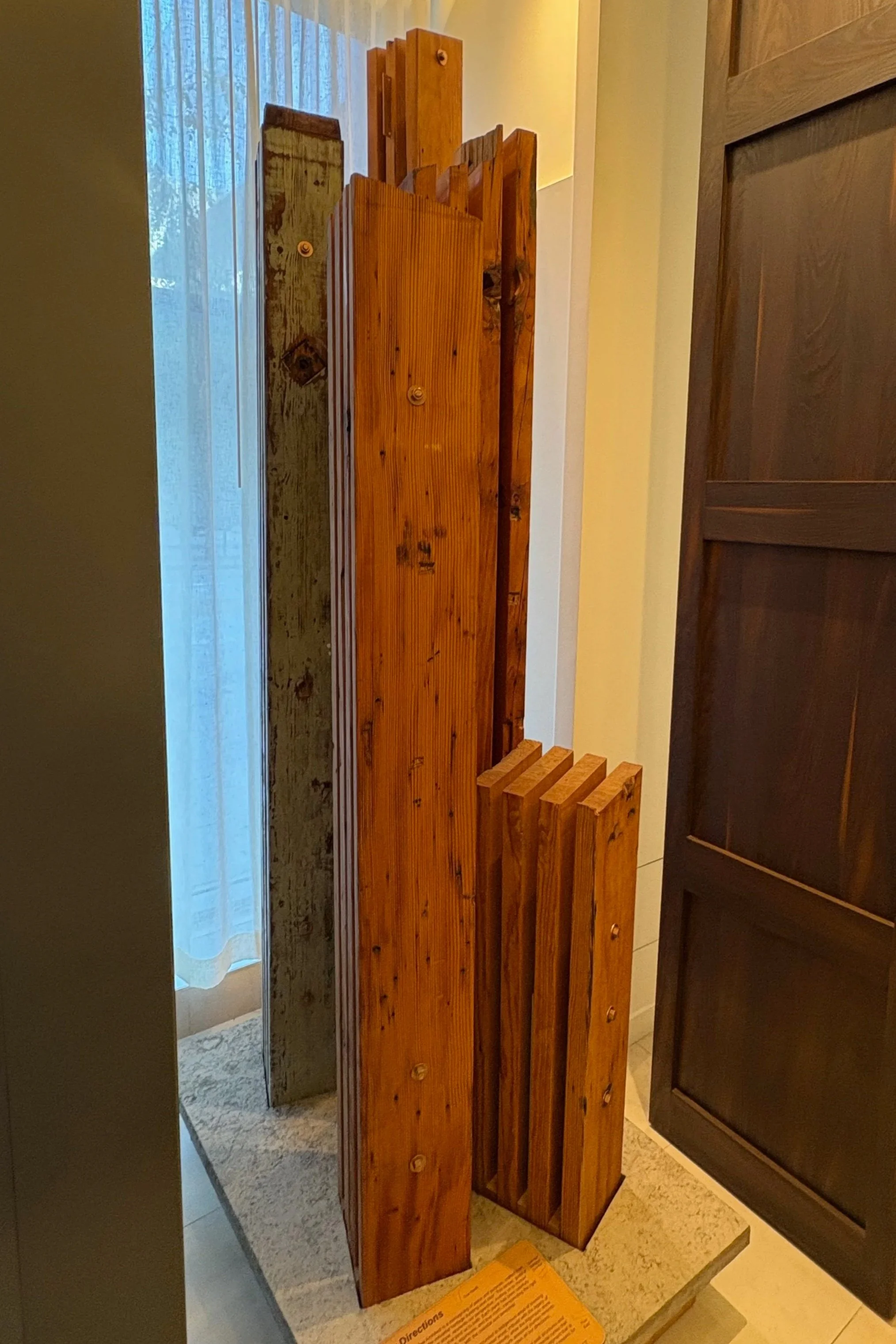 Stacked wooden planks, some stained darker, leaning against a wall, with a window and curtain in the background.