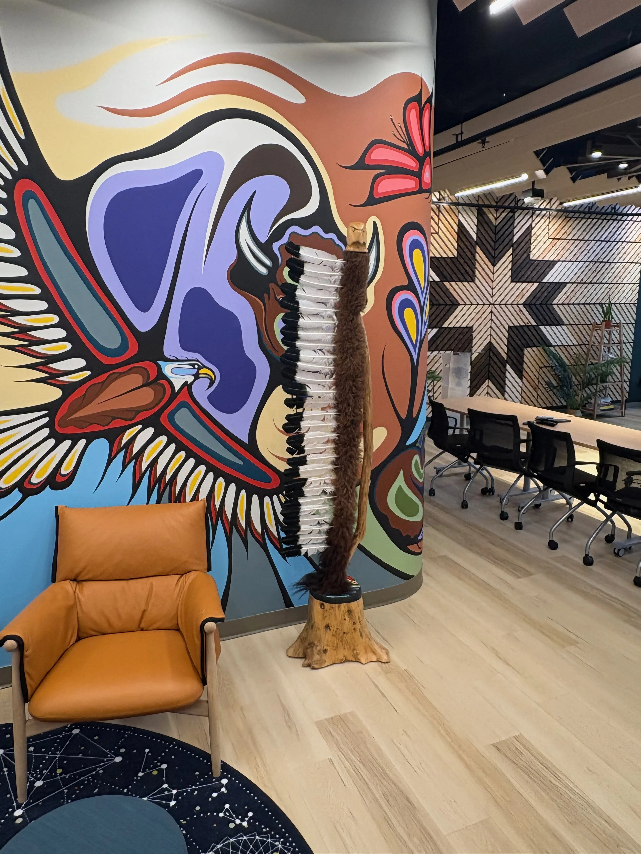 Colorful mural of a bird on the wall with a native American hat decorated with feathers. In front, a leather chair and two fur-covered poles stand next to each other, with a tree stump at the base.