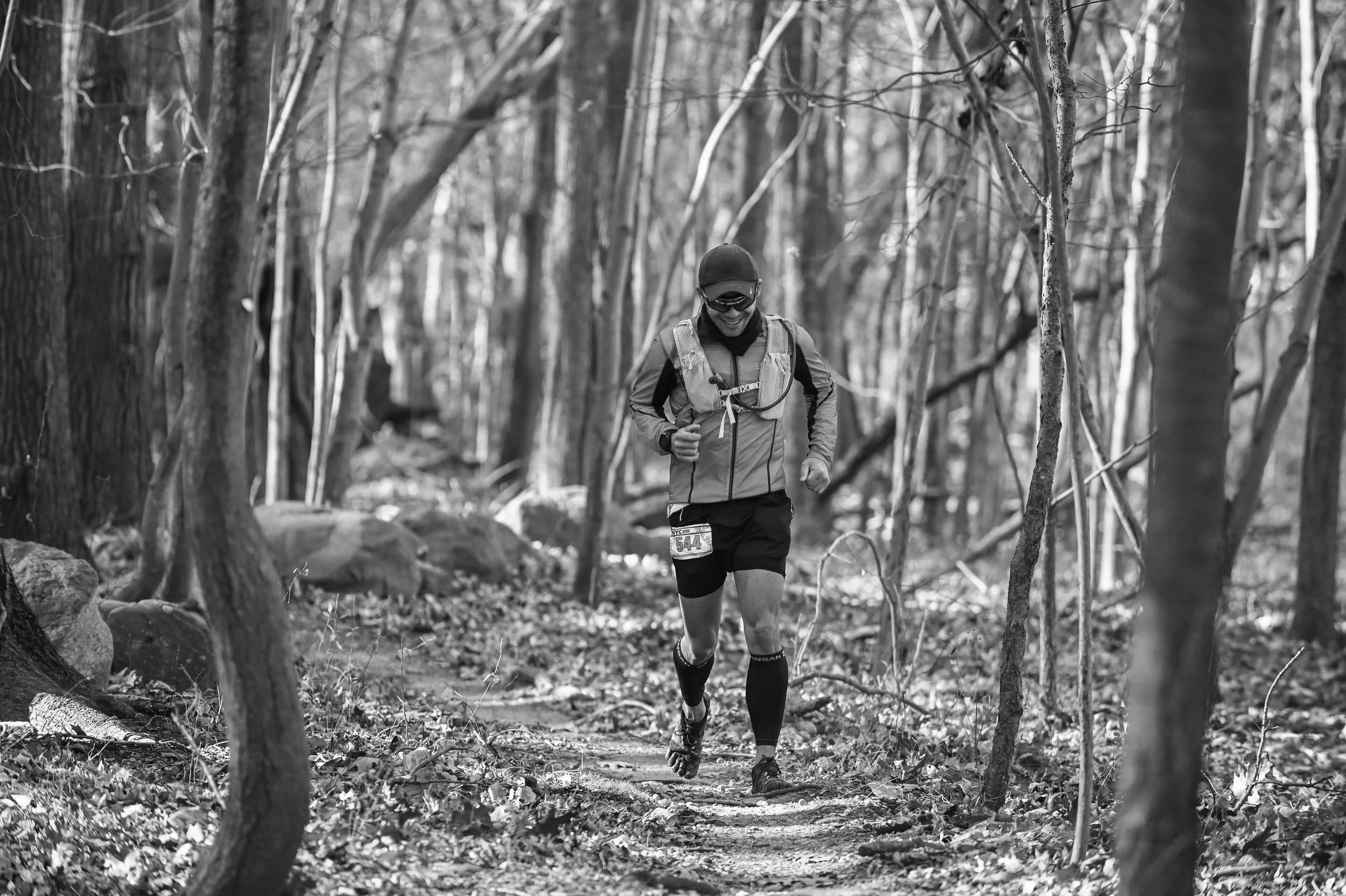 A person running on a trail in a wooded area, wearing a helmet, sunglasses, a jacket, shorts, and a backpack, in black and white.