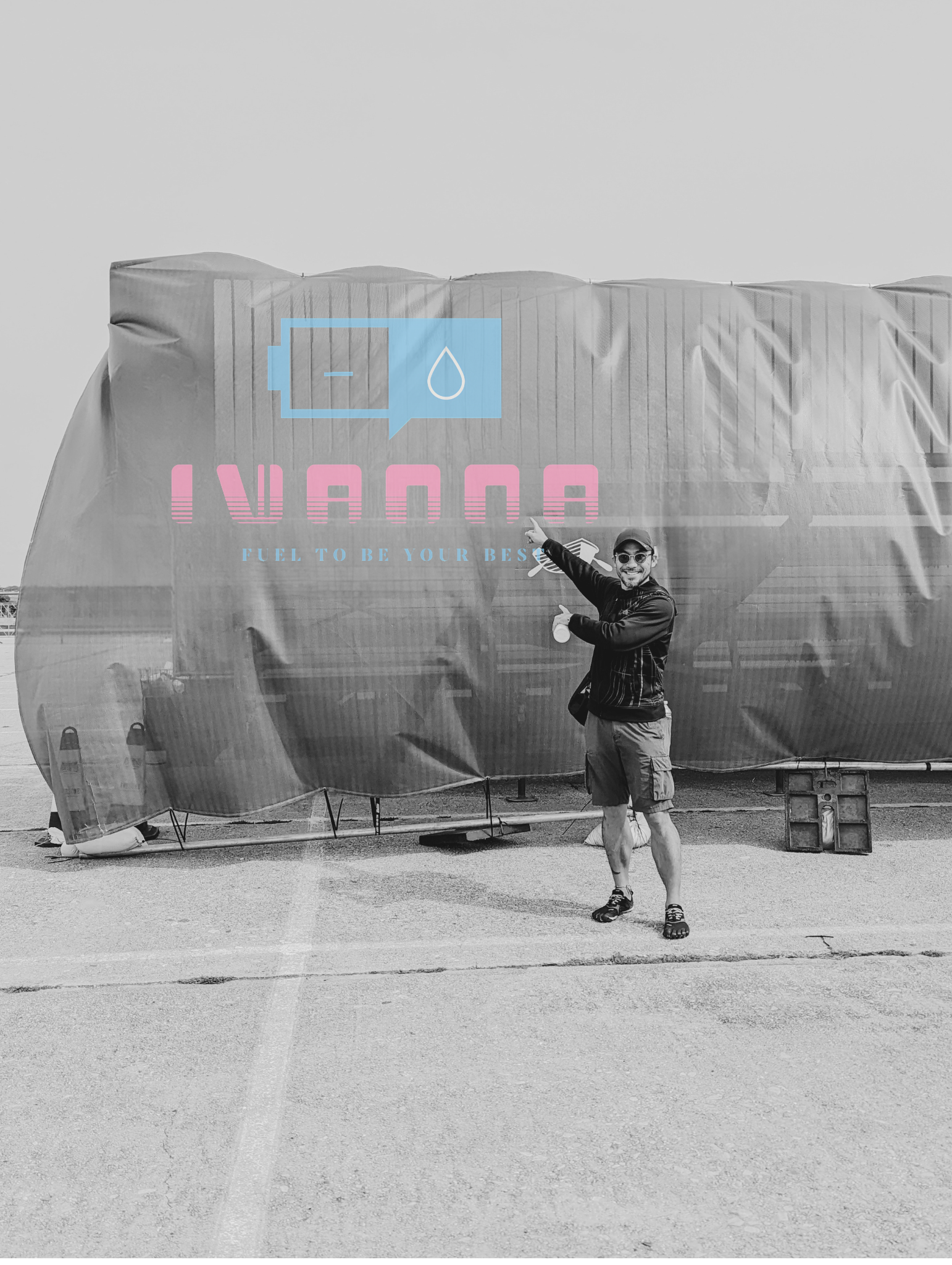 A man standing outdoors on a paved surface, pointing at a large silver tent with a graphic of a battery and water droplet, and the words 'IVANNA' and 'Fuel to be your best' on it.