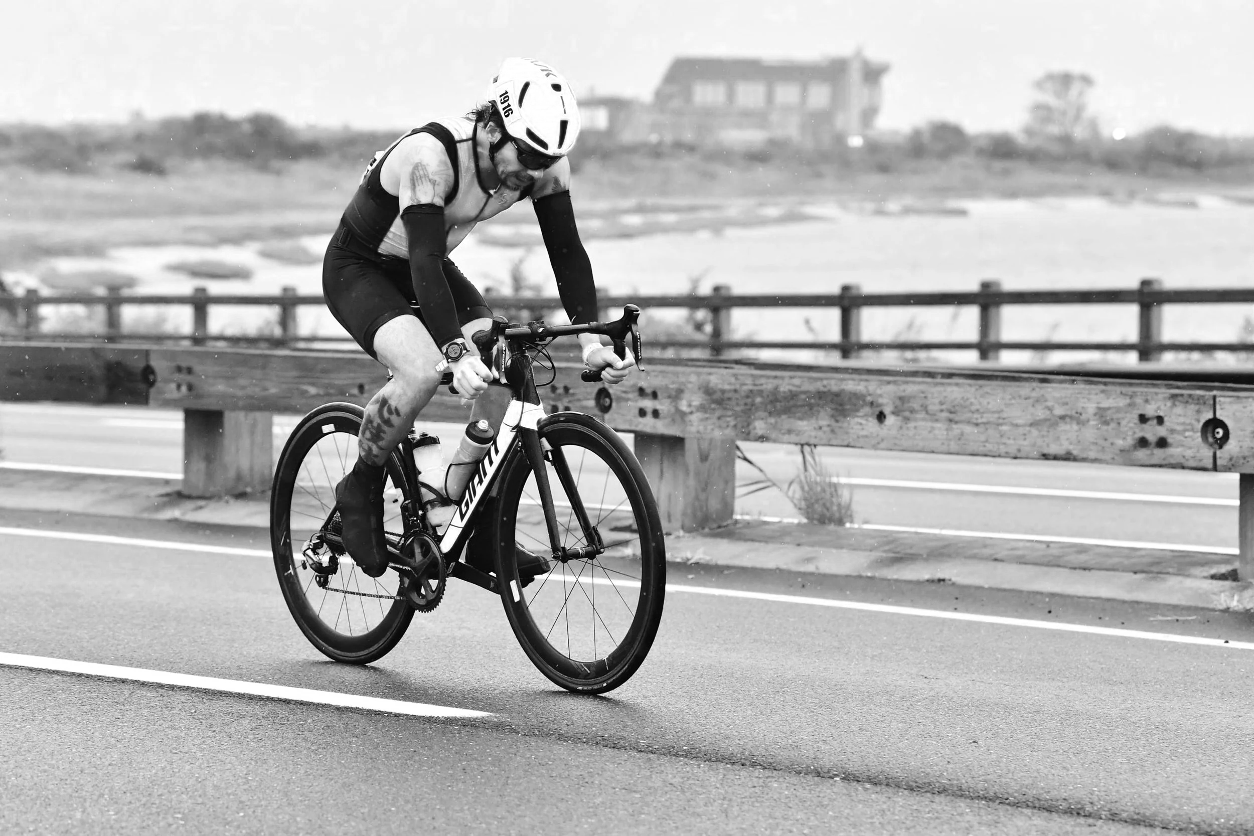 A man in cycling gear riding a bicycle on a road with a blurred background of water and buildings.