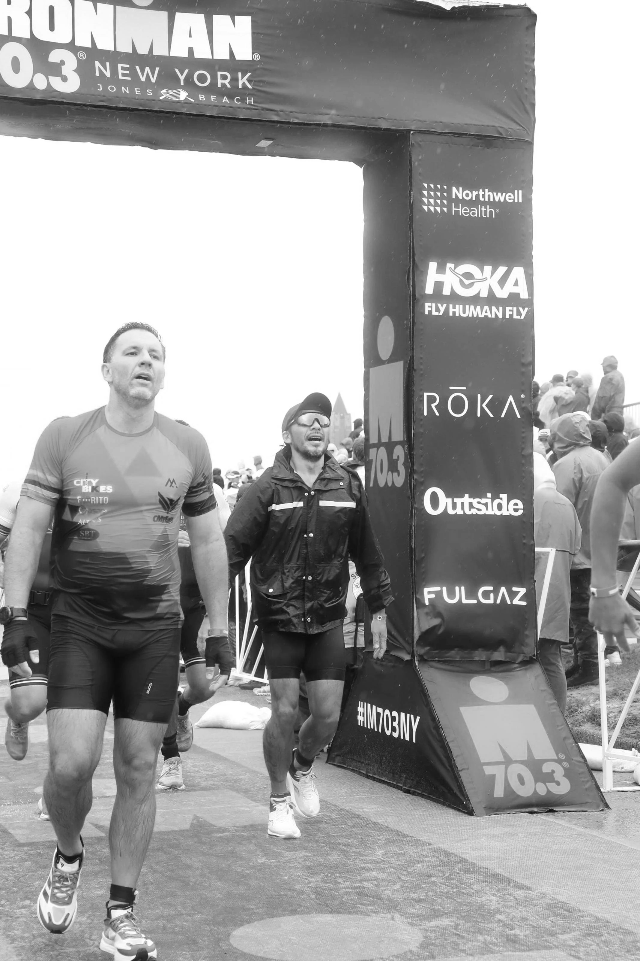 Two male runners participating in a marathon race, crossing the finish line. The background shows a crowd of spectators and various race banners.