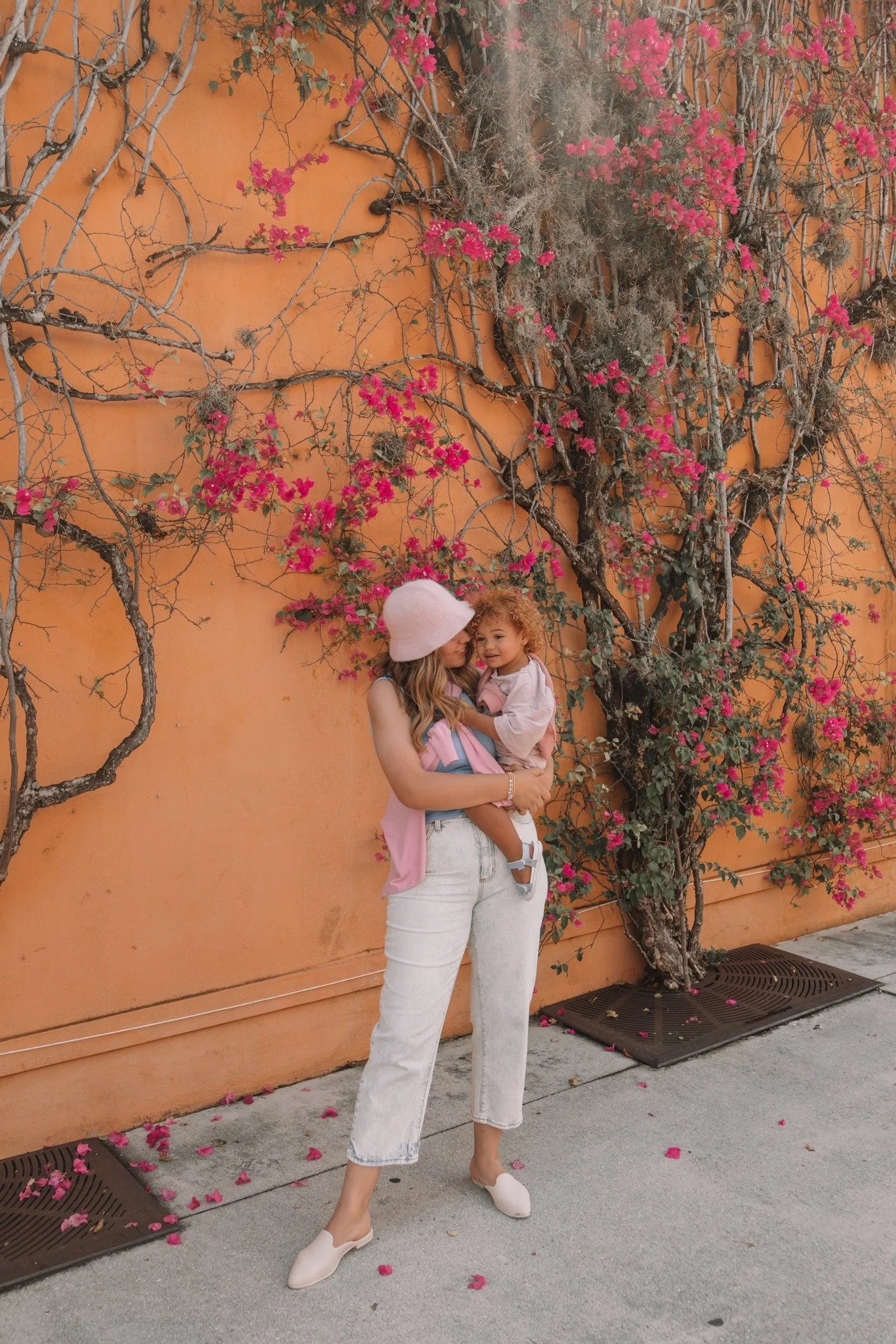 Woman holding her toddler daughter while posing in front of an orange stucco wall.