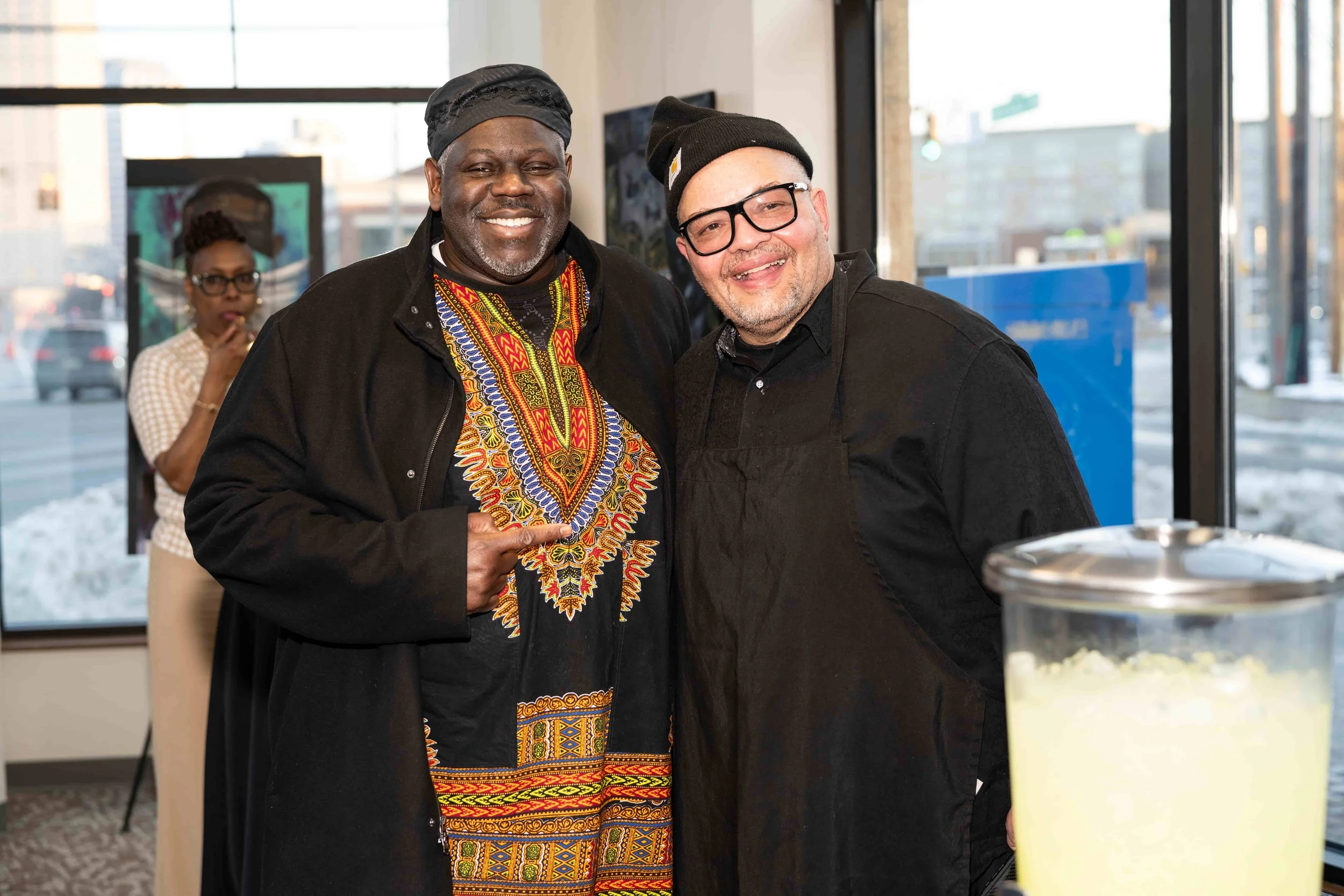 Two men smiling and posing together indoors, one wearing a black jacket and colorful African-style shirt, the other wearing a black beanie, glasses, and a black shirt and apron. A woman is visible in the background, standing near a window.