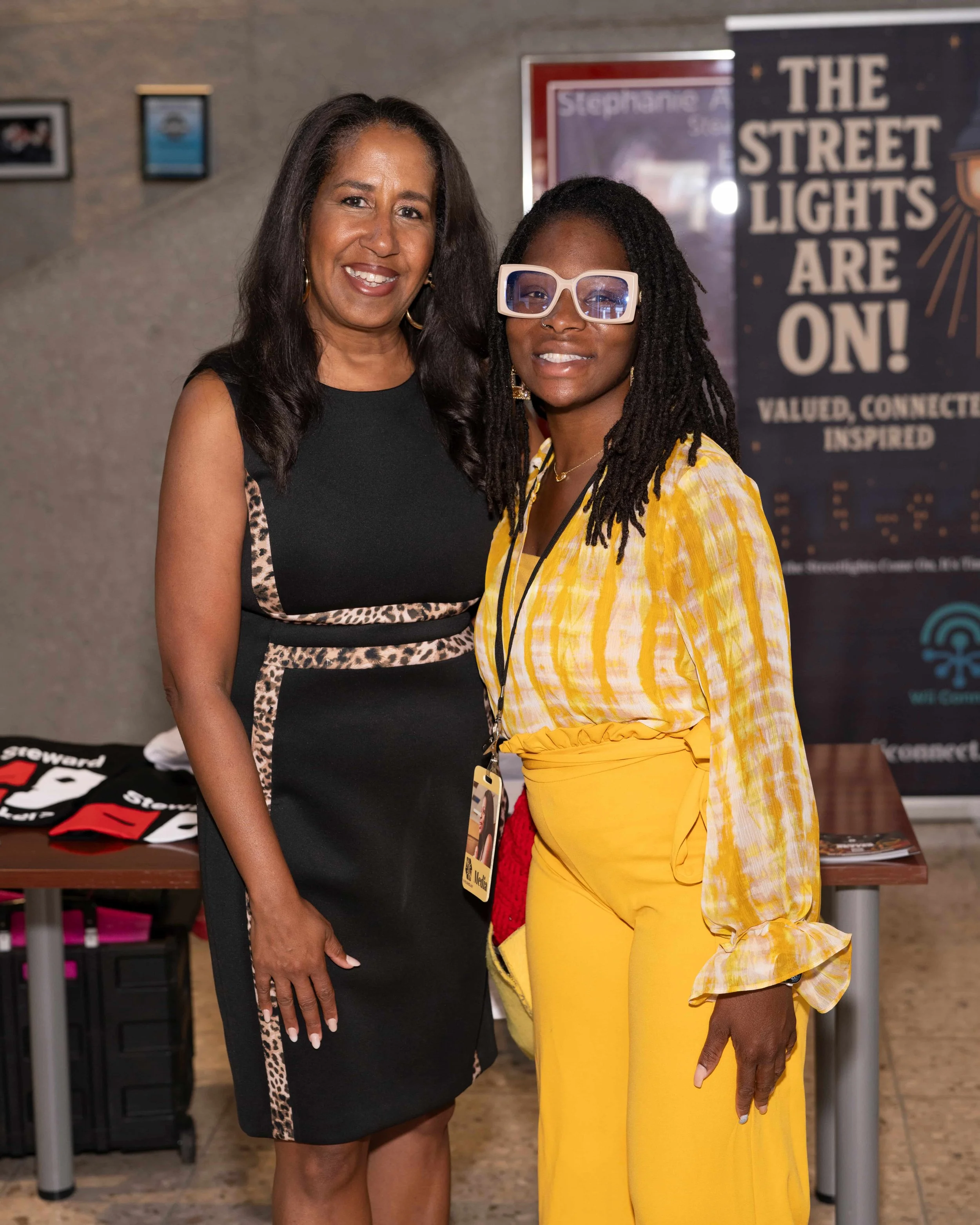 Two women posing for a photo indoors, one wearing a black dress with leopard print accents and the other in a yellow and white patterned blouse with yellow pants, standing in front of a poster that reads 'The Street Lights Are On!'