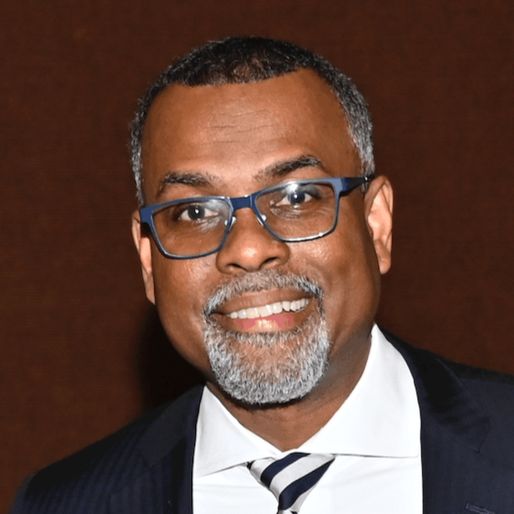 A smiling man with dark skin, glasses, short gray hair, and a gray beard, wearing a dark suit, white shirt, and striped tie, against a brown background.