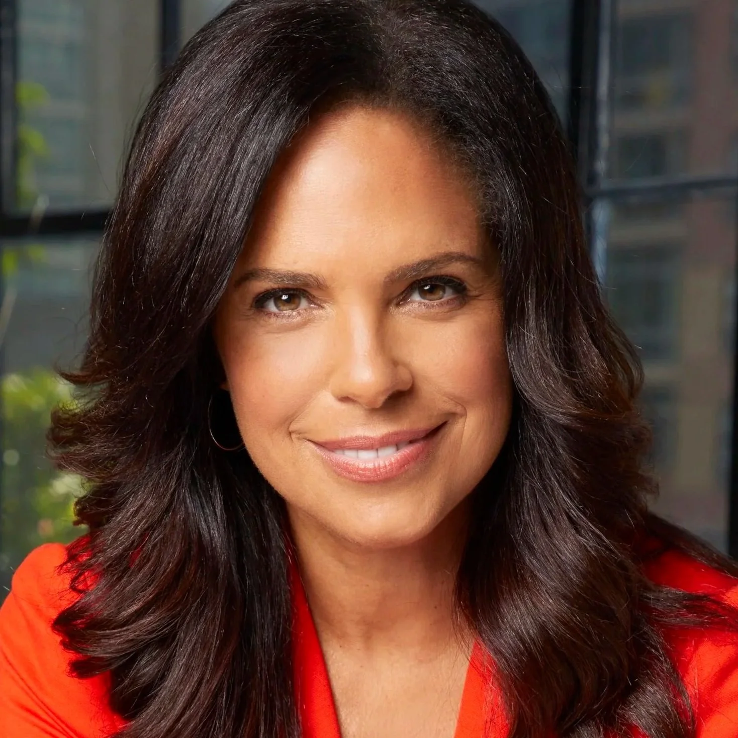 A woman with long dark brown hair, wearing a red top, smiling, in front of a window with an urban cityscape background.