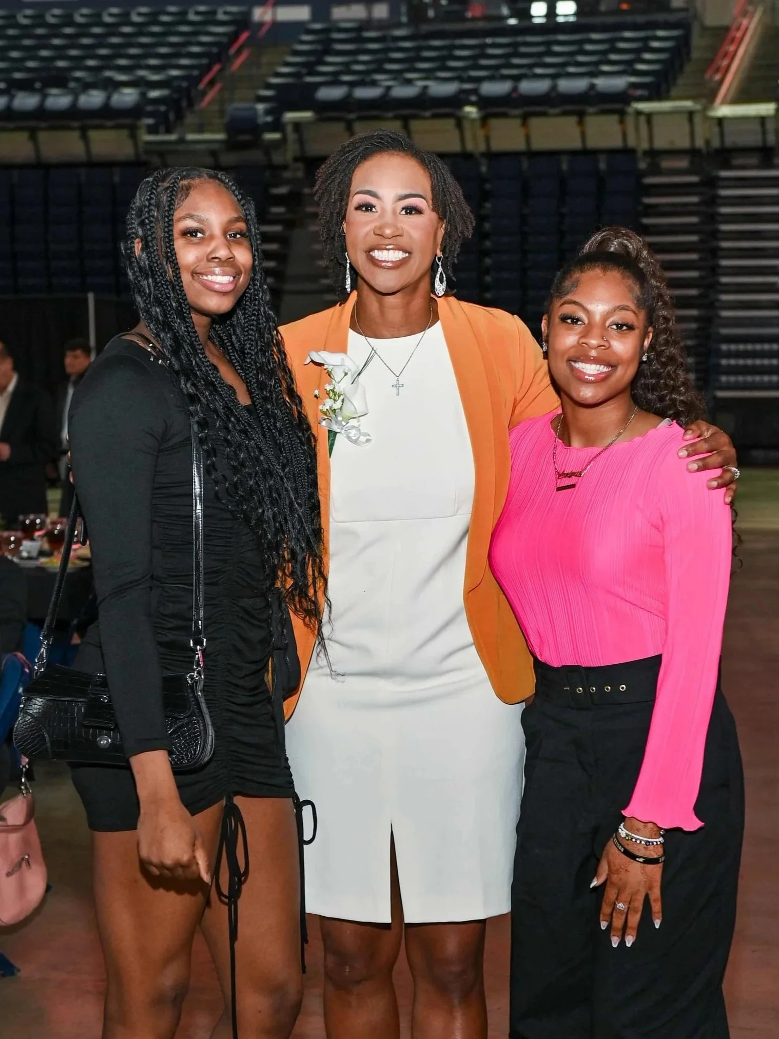 Three women standing together smiling at an indoor event, with one woman in a white dress and orange blazer in the center, and two women in black and pink outfits on either side.