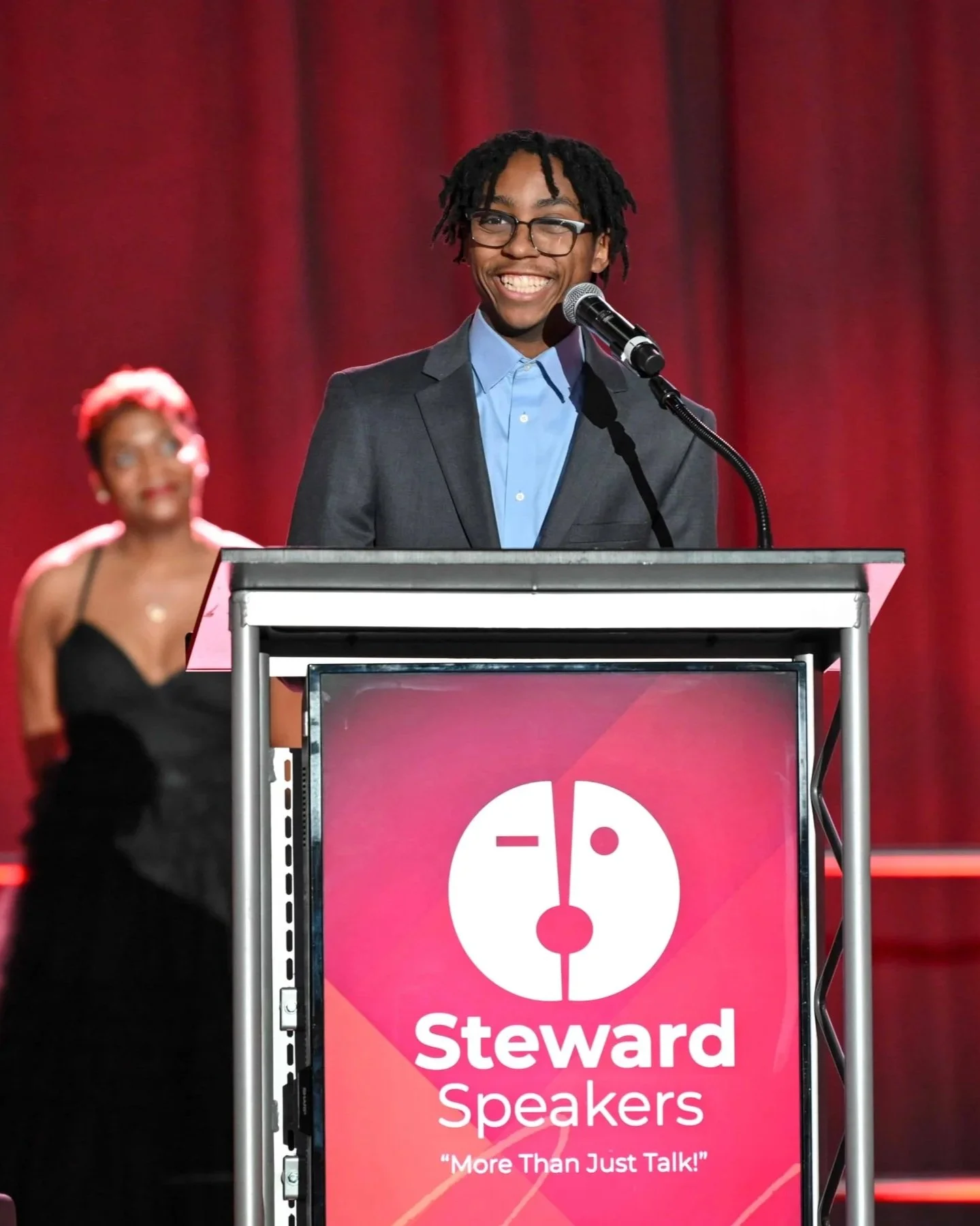 A young man with glasses and dreadlocks, dressed in a suit, is speaking at a podium with a microphone. There is a woman in a black dress in the background, and a sign on the podium reads 'Steward Speakers' and includes a logo of a face with makeup and the slogan 'More Than Just Talk!'