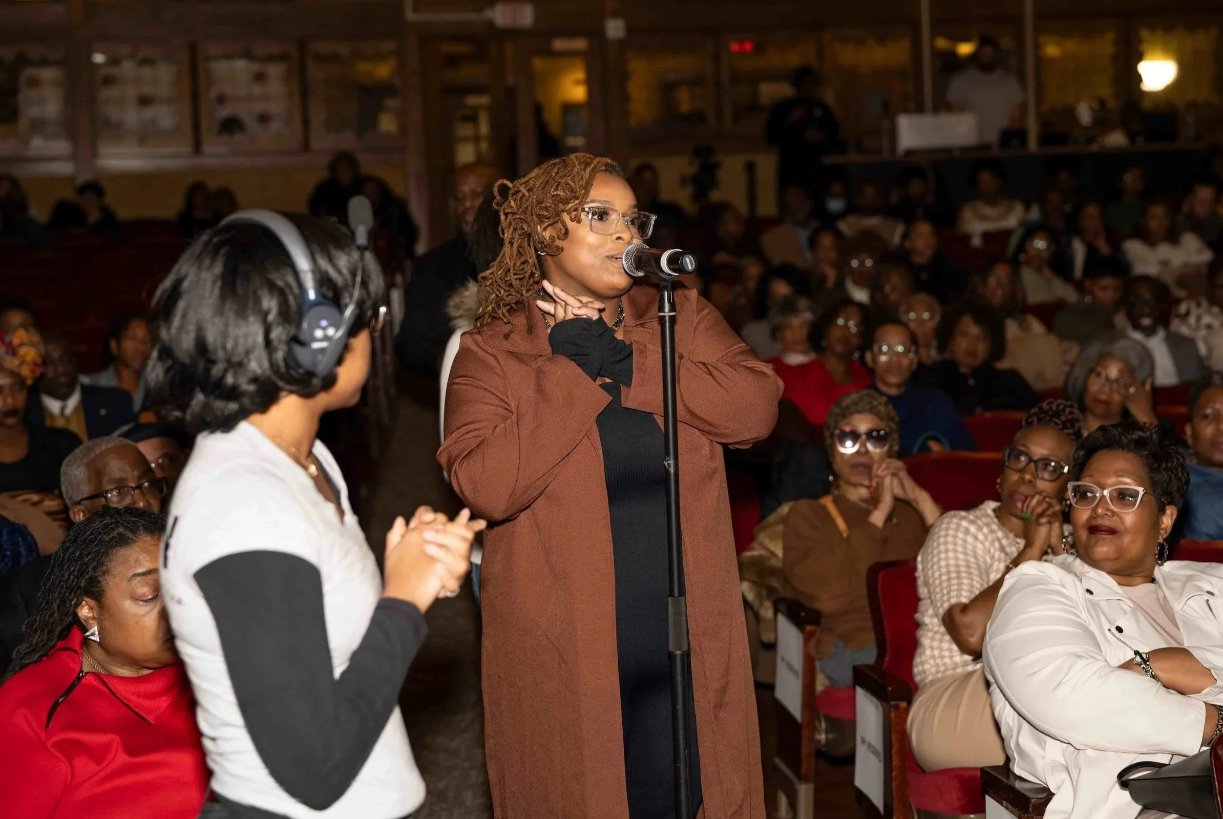 A woman with glasses and dreadlocks speaking into a microphone at an indoor event, surrounded by seated audience members.