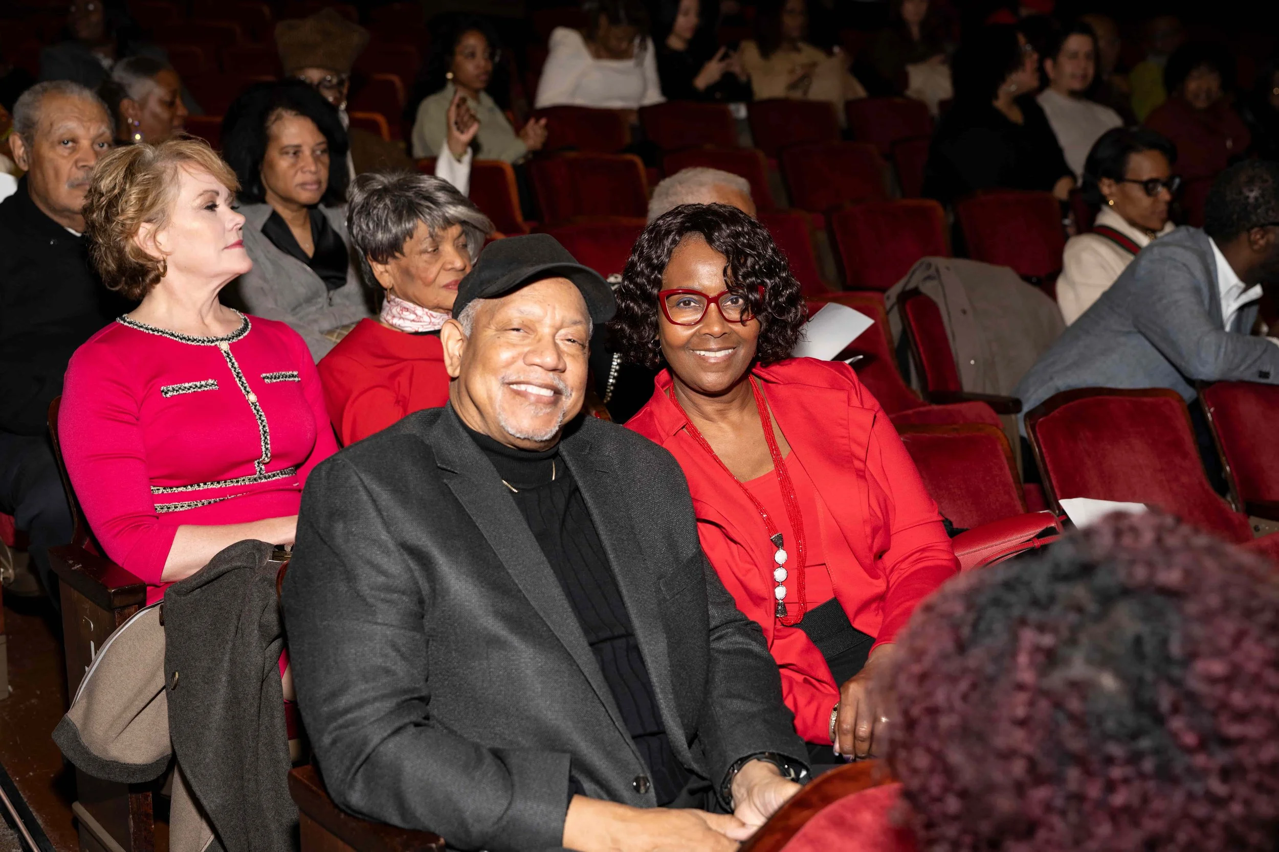 People sitting in an auditorium, with two individuals in the front row smiling at the camera. The man wears a black blazer and cap, and the woman wears glasses and a red top. Other audience members sit behind them.