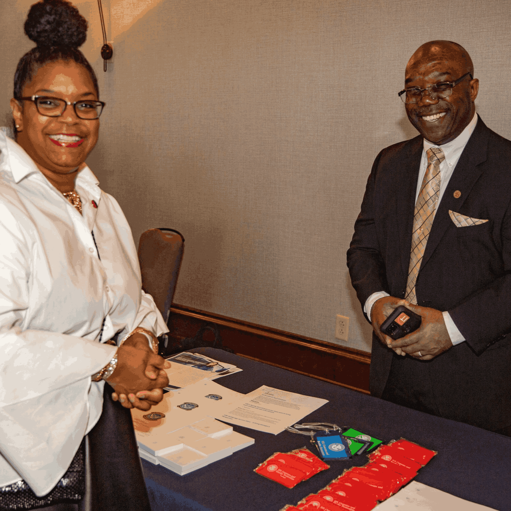 A woman and a man standing at a conference table with informational materials, badges, and red lanyards, smiling and posing for the photo in a professional setting.