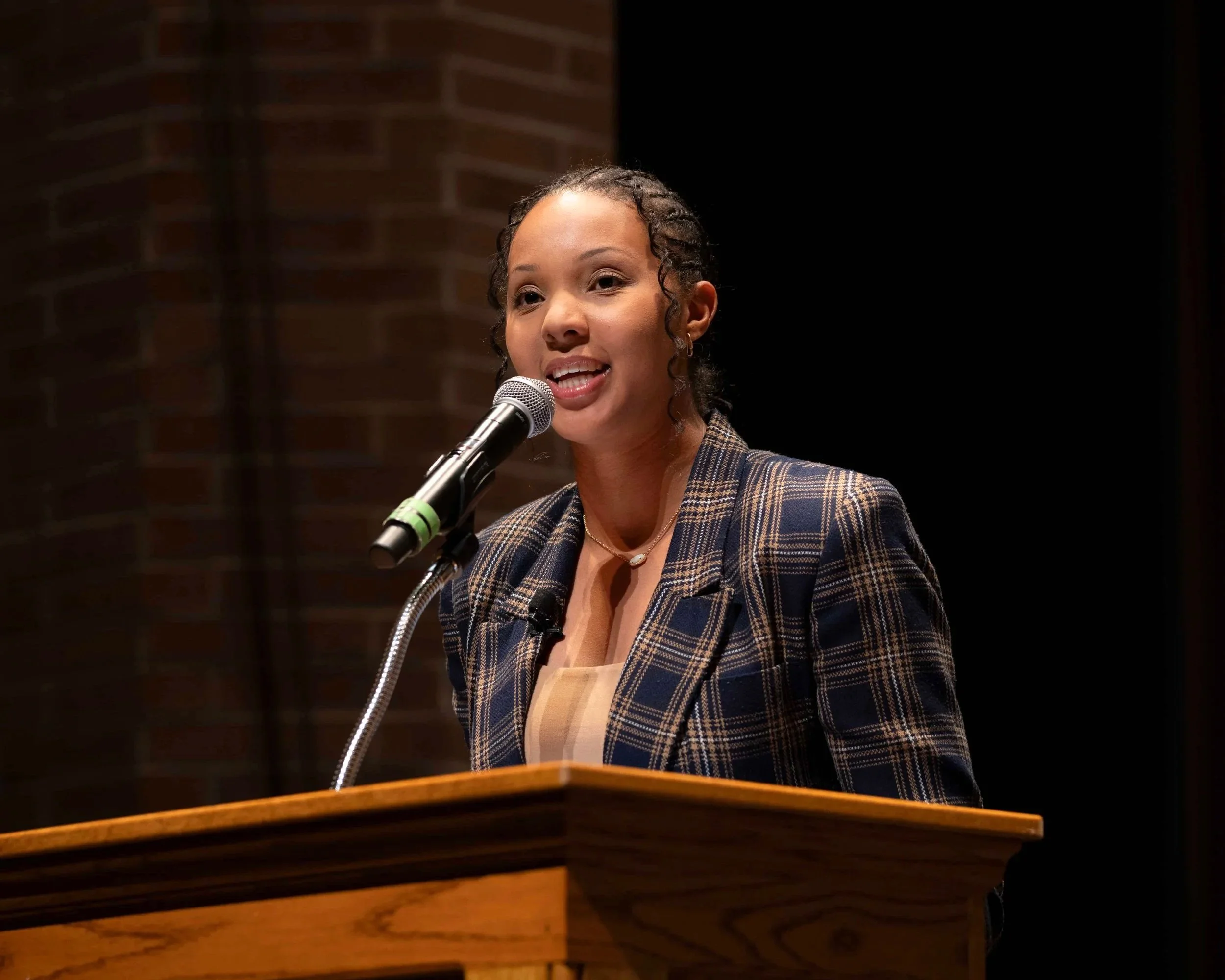 A woman in a plaid blazer speaks into a microphone at a podium.