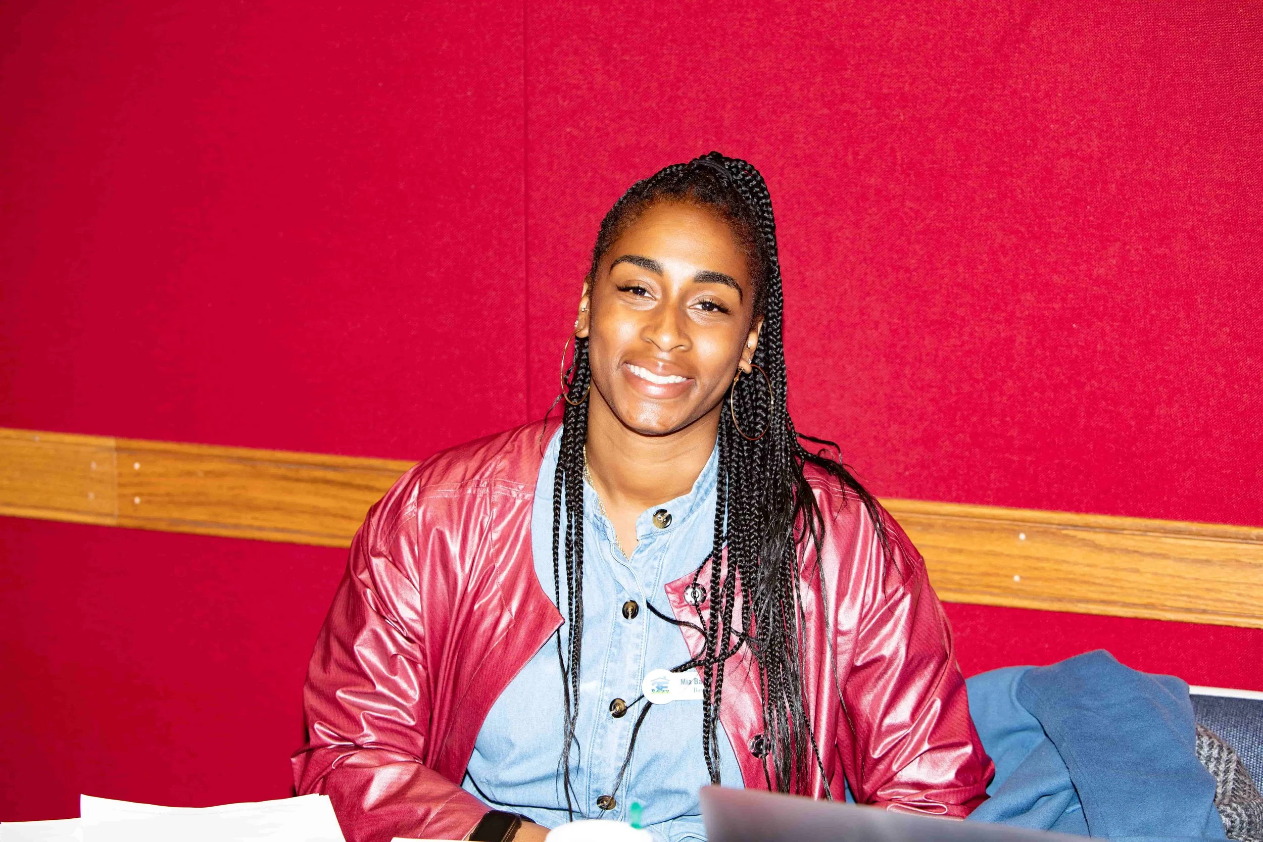 A woman with braids smiling, wearing a red jacket and light blue shirt, sitting at a table with papers and a laptop, against a red wall.