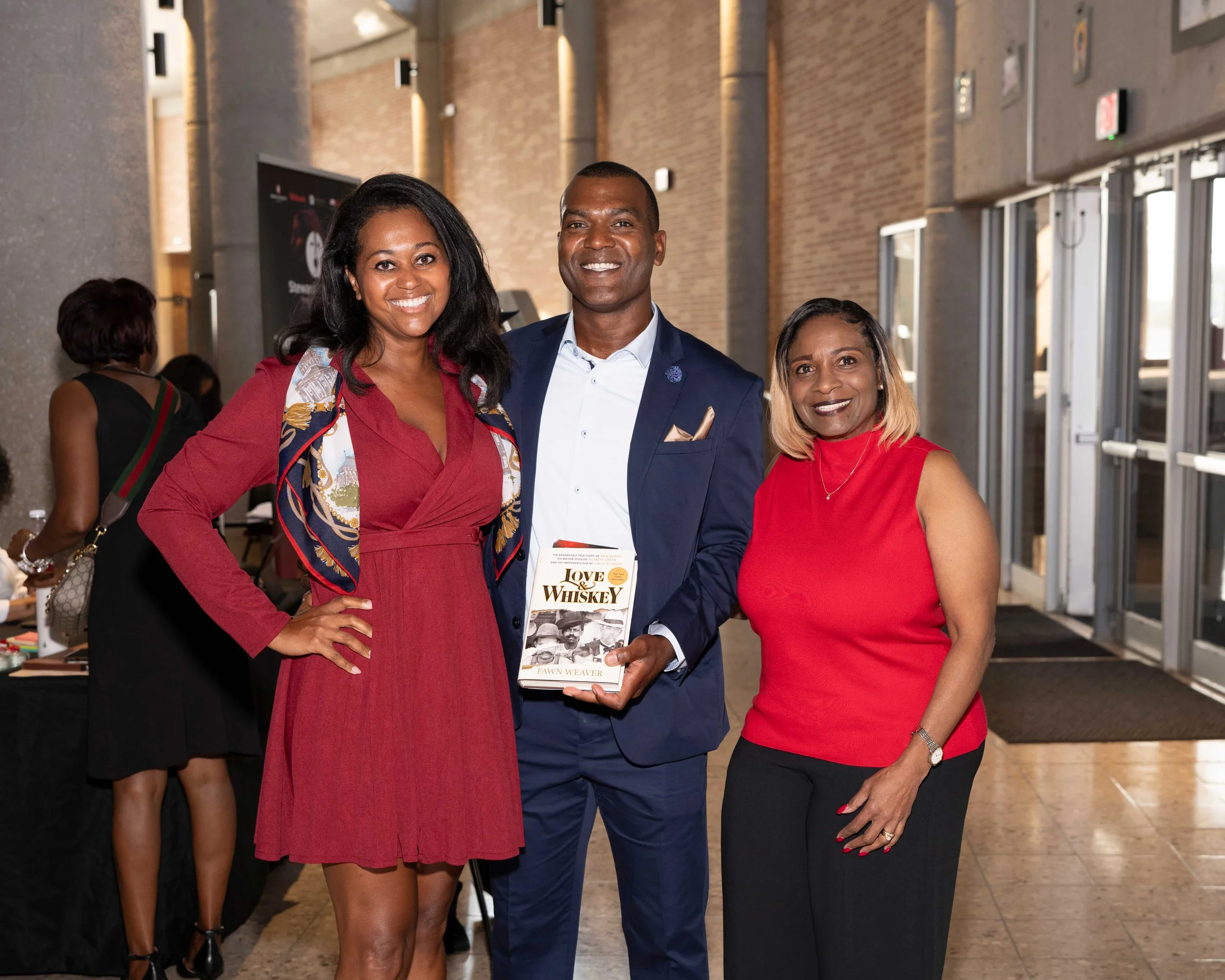 Three people, two women and one man, standing together smiling at an indoor event. The man is holding a book titled "Love & Whiskey" by Fawn Weaver. The woman on the left is wearing a red dress with a colorful scarf wrapped around her shoulders, and the woman on the right is dressed in a sleeveless red top and black pants.