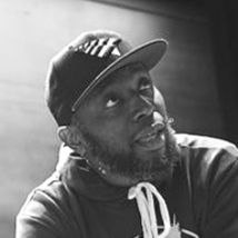 Black and white photo of a man wearing a cap, with a beard and mustache, looking at the camera.