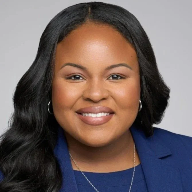 A woman with long black hair, smiling, wearing a blue blazer and a silver necklace against a gray background.