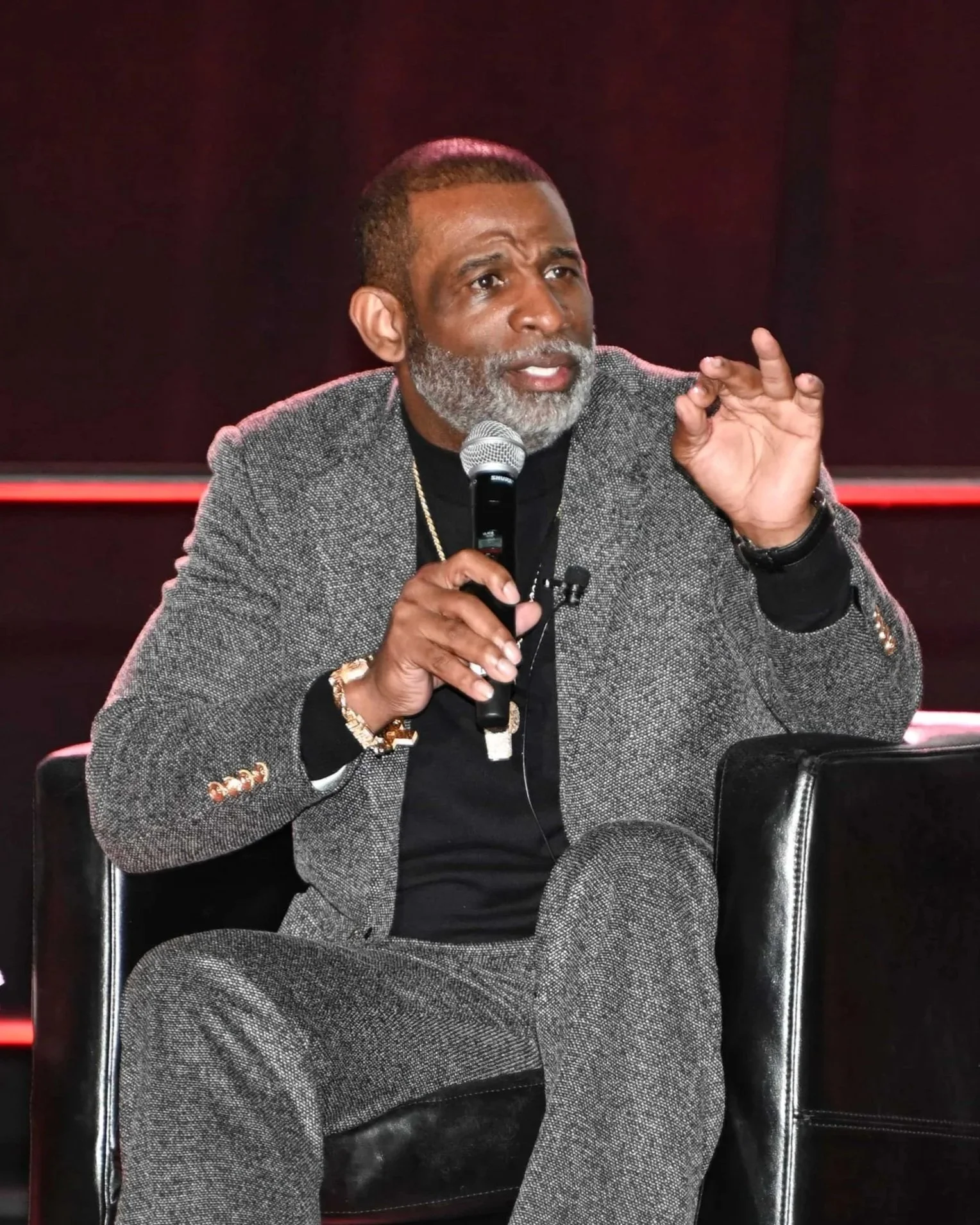 A man with gray hair and beard, wearing a gray blazer and black shirt, sitting on a black chair, holding a microphone, and gesturing with his right hand.