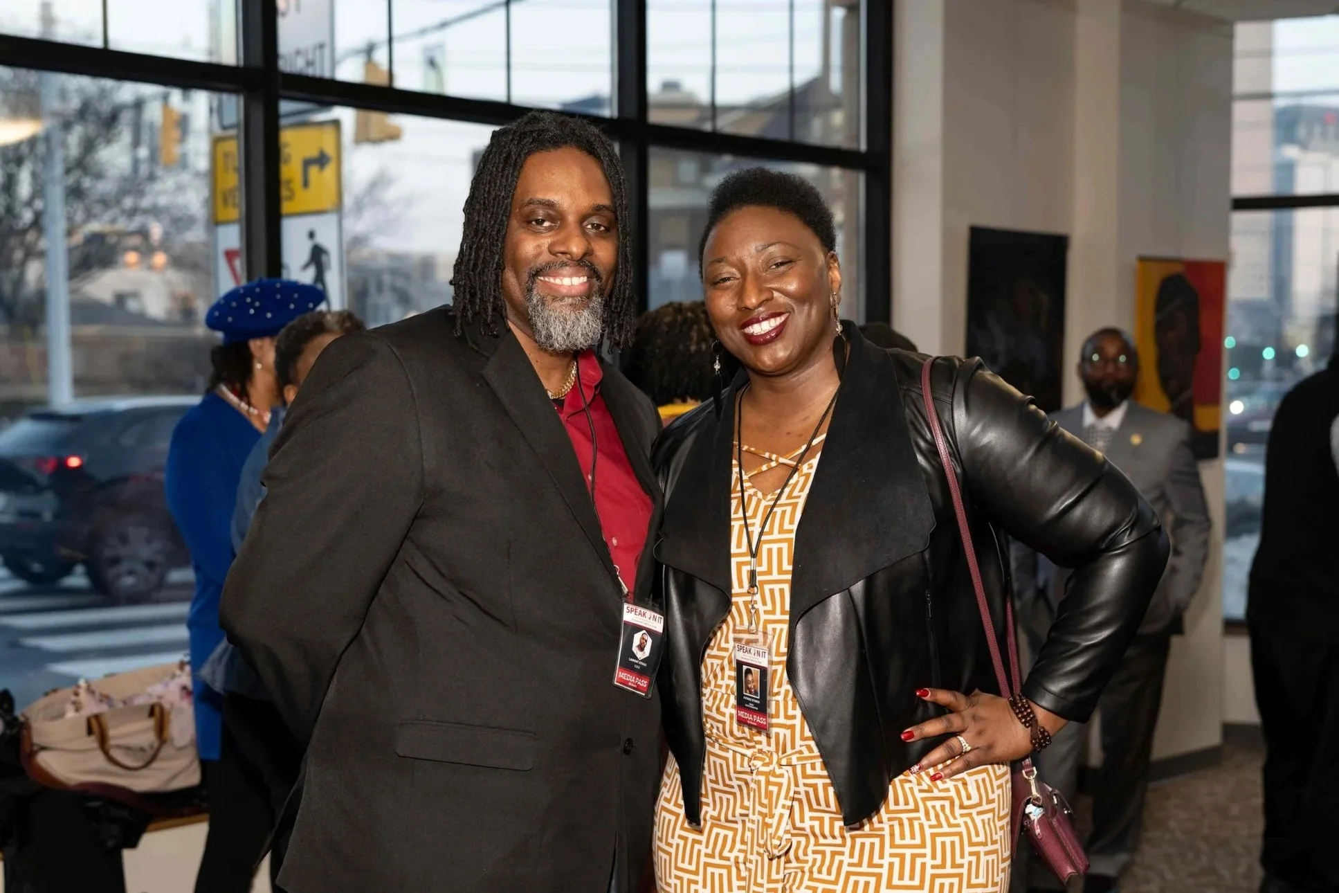 A man and a woman smiling and posing together indoors during an event, with badges around their necks, others in the background, and large windows showing a city street outside.