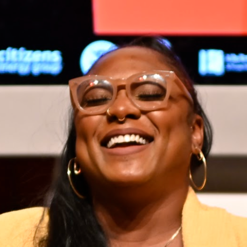 A woman with glasses and hoop earrings smiling in an indoor setting with a blurred background containing bright lights and signs.